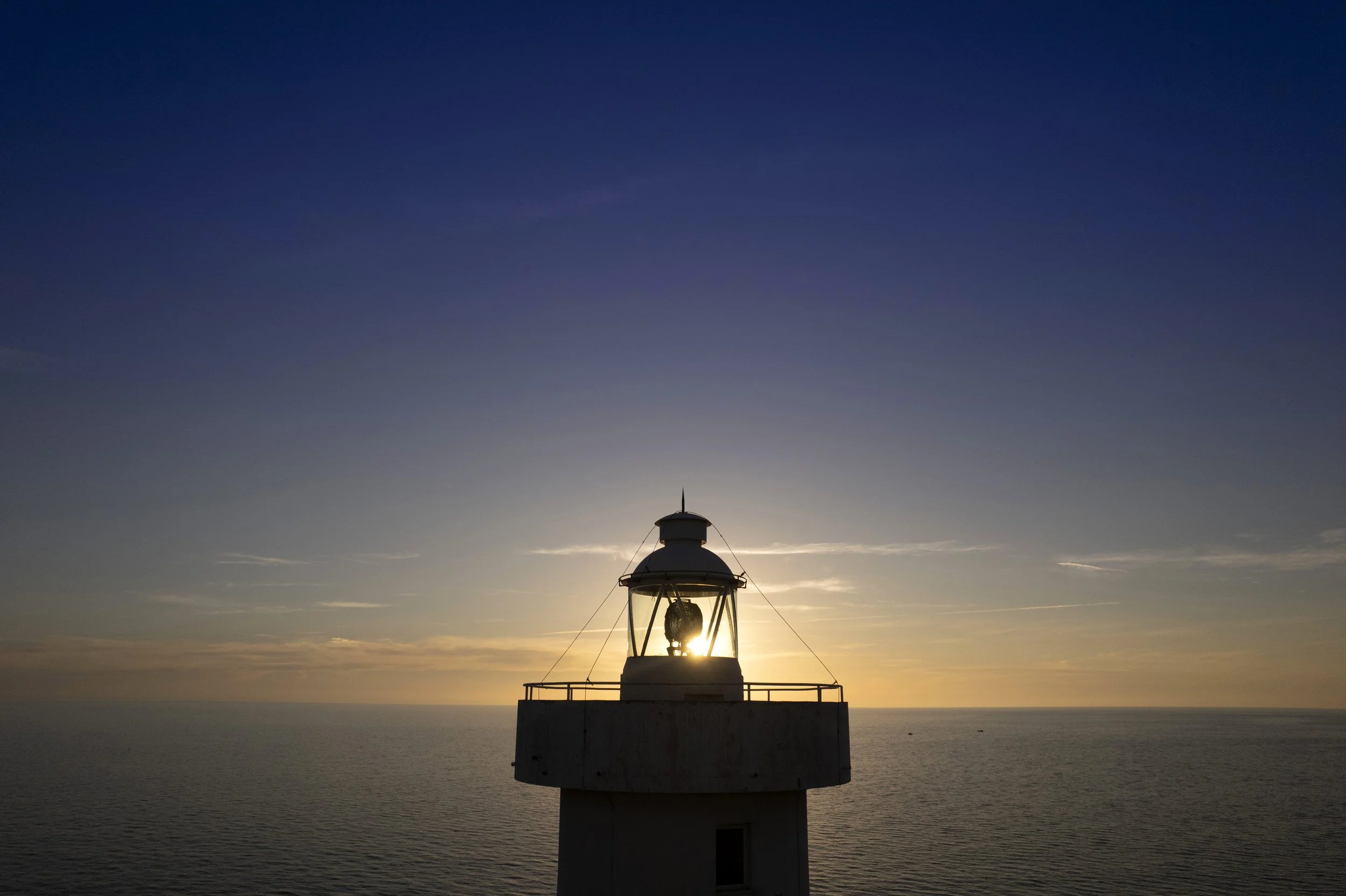 Lighthouse at sunset over the ocean with a clear sky and the sun setting on the horizon.