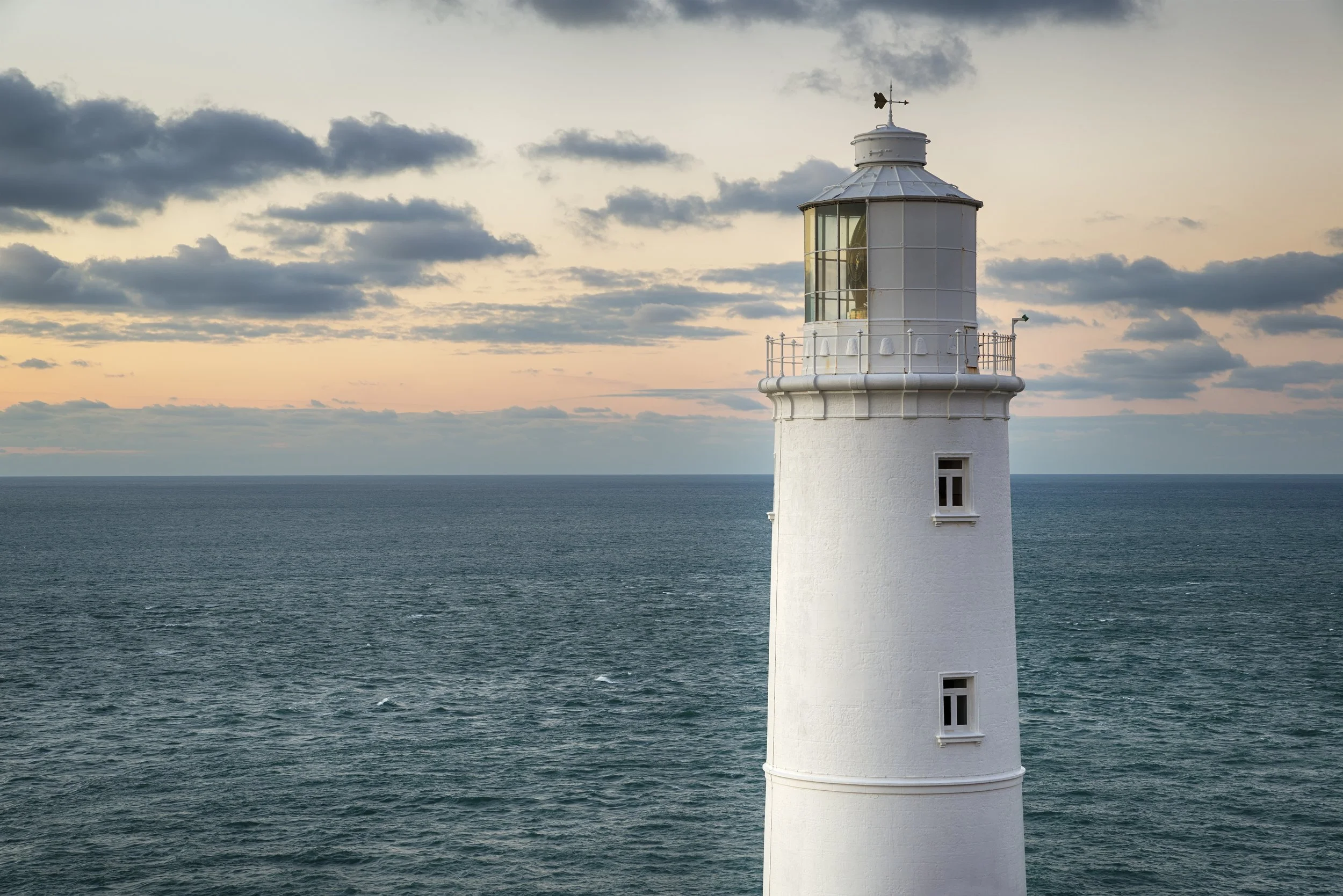 A white lighthouse standing on the edge of the sea during sunset with cloudy skies.