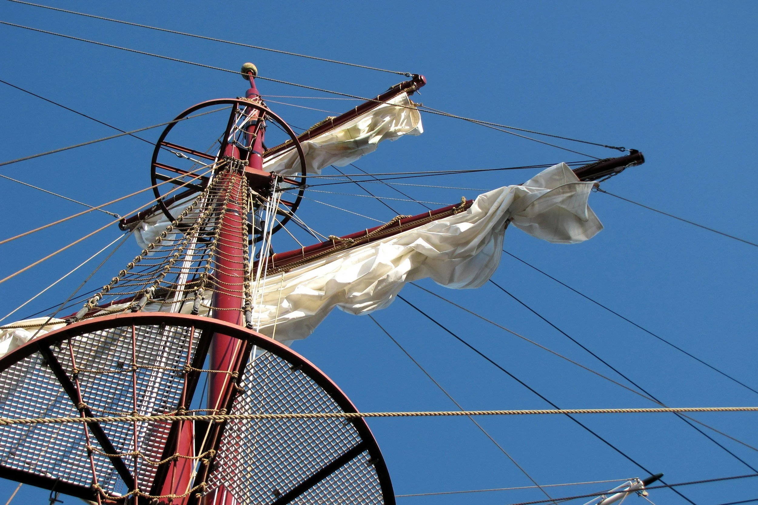 View of the top of a historic sailing ship's mast with white sails furled, against a clear blue sky.