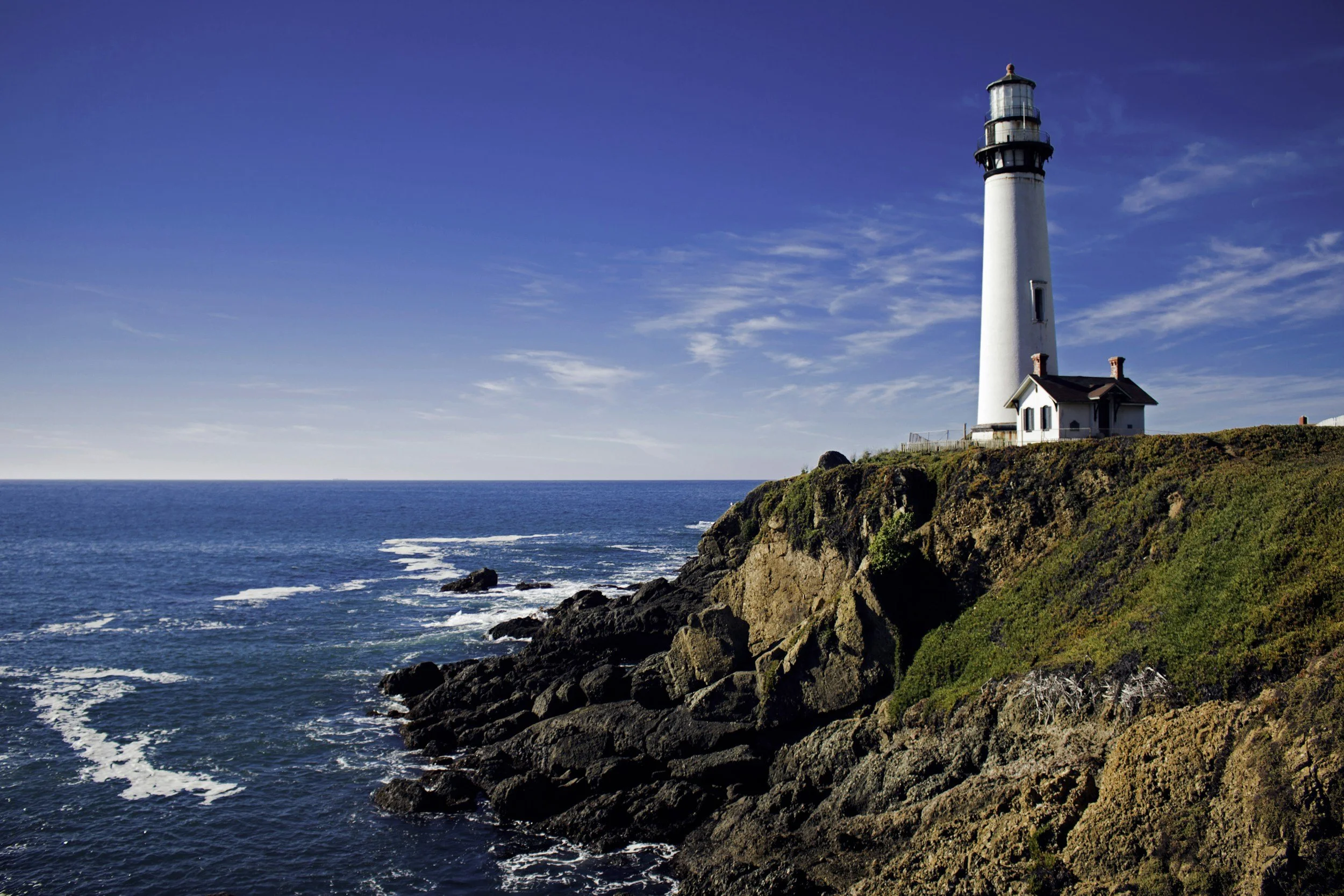 A lighthouse on a rocky cliff overlooks the ocean against a clear blue sky.