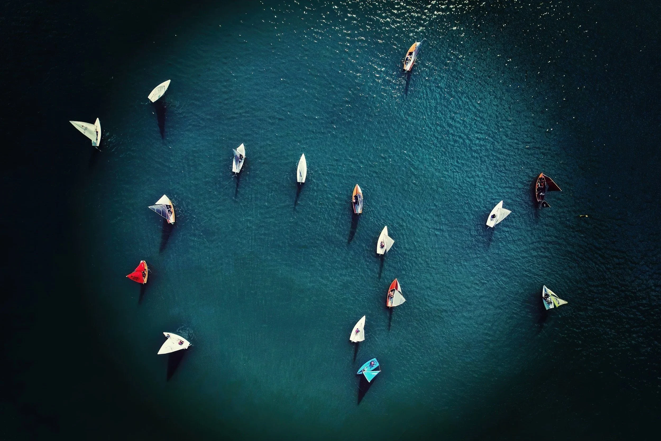 Aerial view of multiple sailboats cruising on calm ocean water, with their shadows visible on the surface.