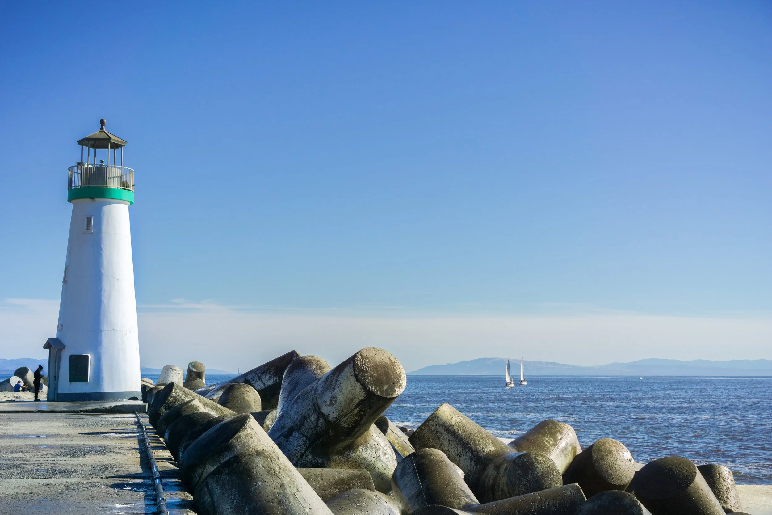 A lighthouse on a pier by the ocean with sailboats in the distance and large rocks in the foreground.