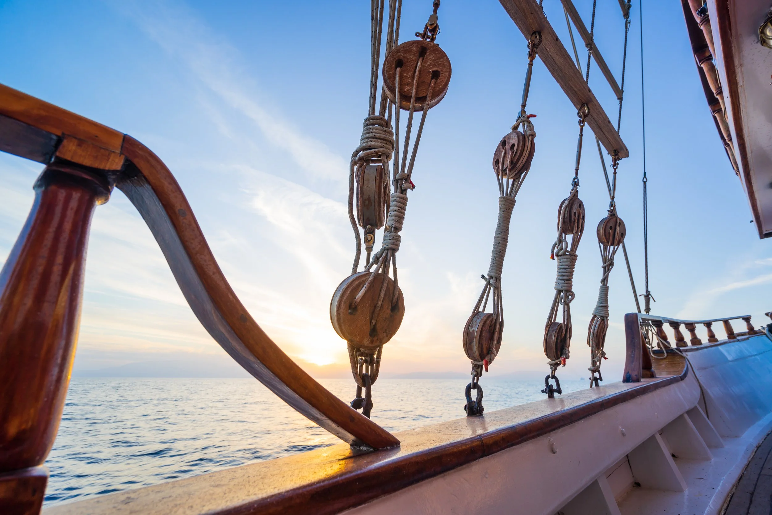View from a wooden ship's deck showing the rigging and ropes against a blue sky with a setting sun over the ocean.