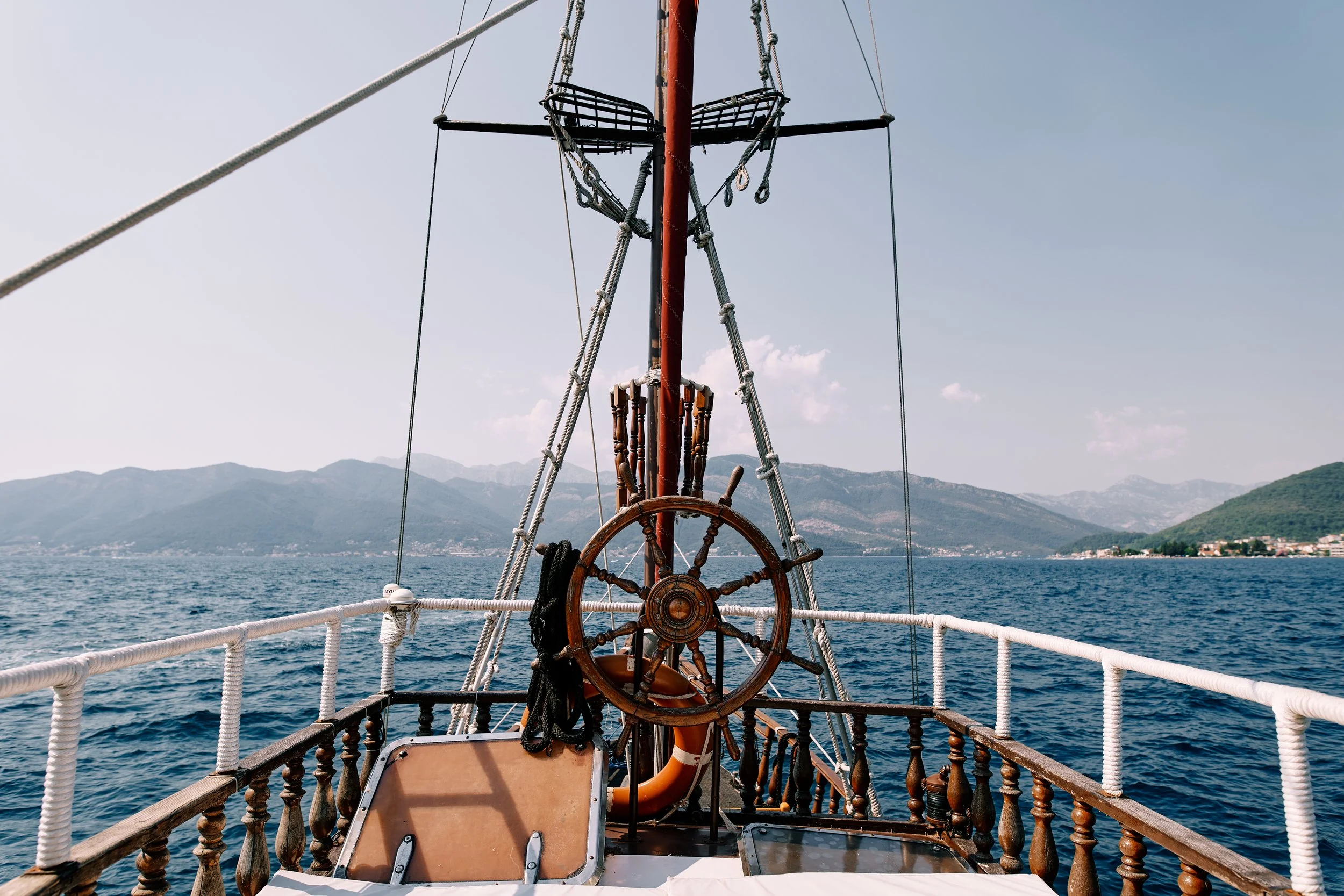 View from the deck of a sailboat showing the steering wheel, ropes, and the boat's bow with a scenic ocean and mountains in the background.