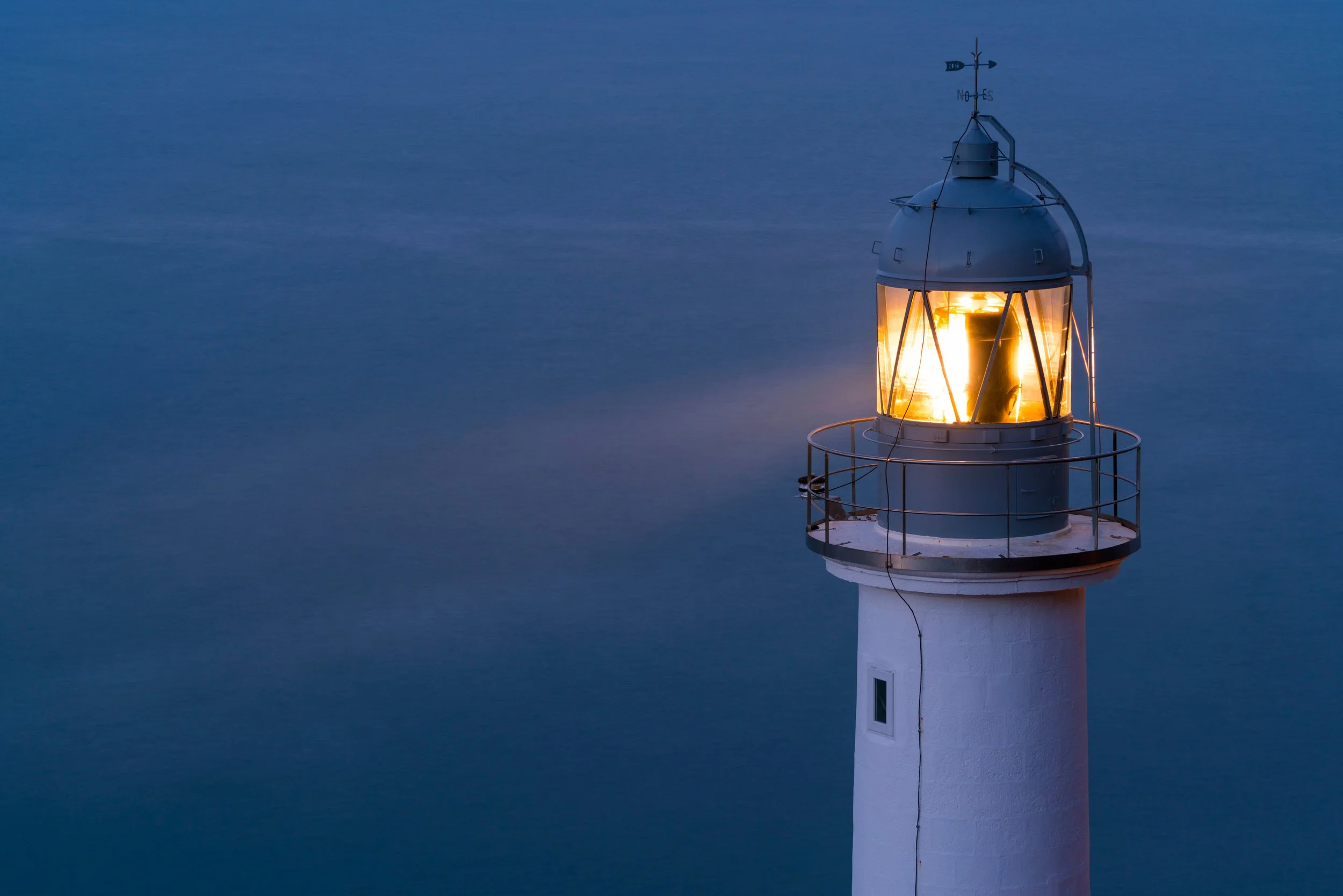 A lighthouse with a glowing light at dusk or dawn, surrounded by calm waters.