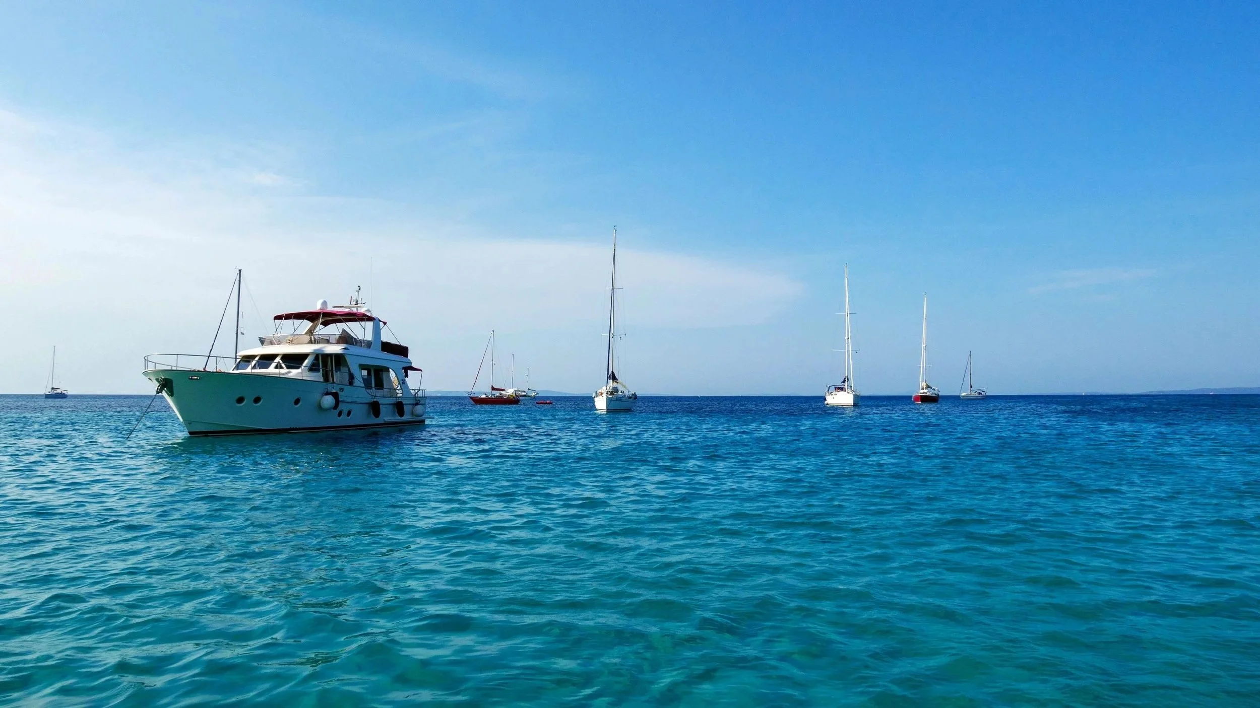 Several boats and yachts floating on calm blue water on a clear day with a bright sky.