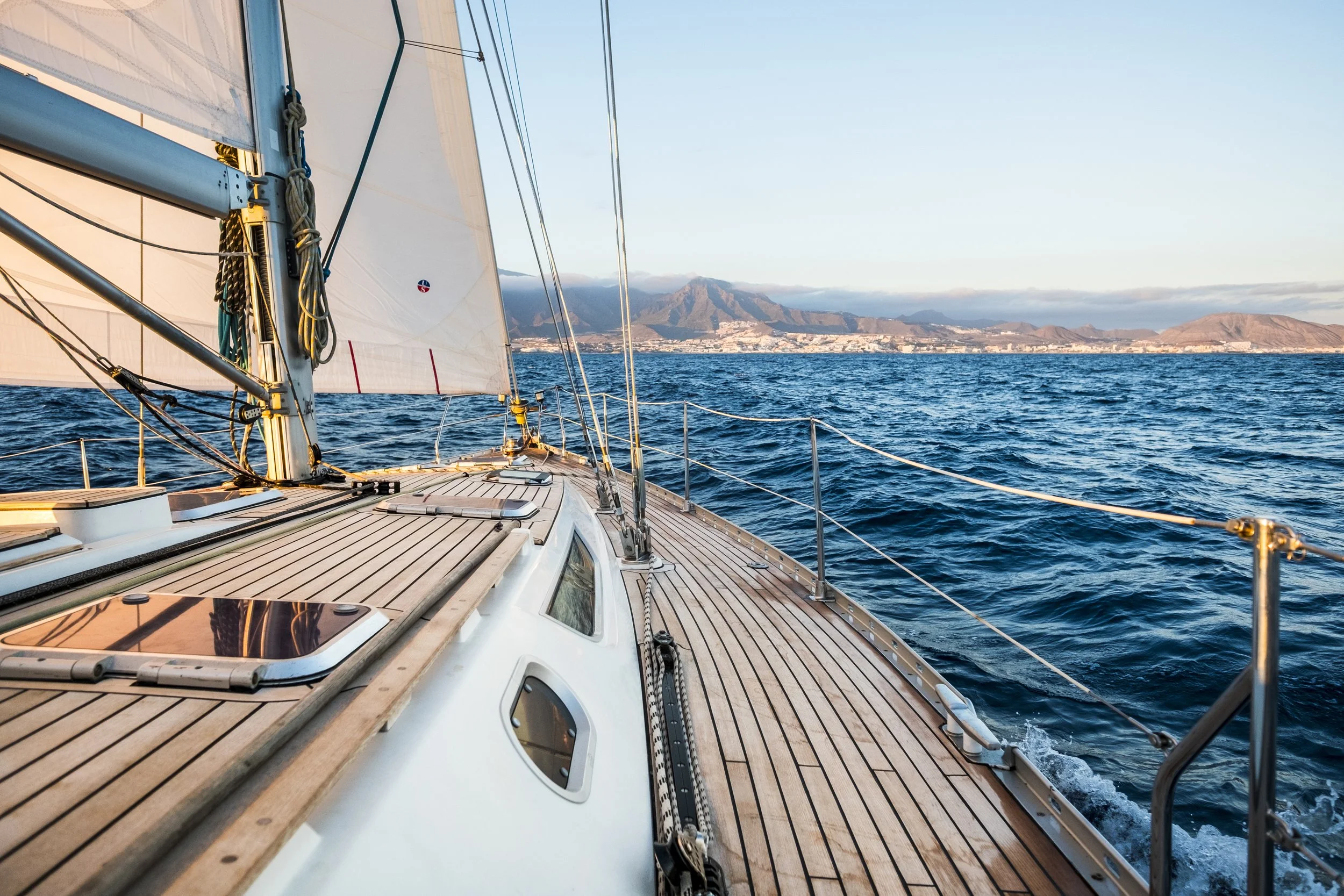 View from the deck of a sailboat, showing the wooden deck, ropes, and sails, with a scenic ocean landscape and distant mountains in the background.