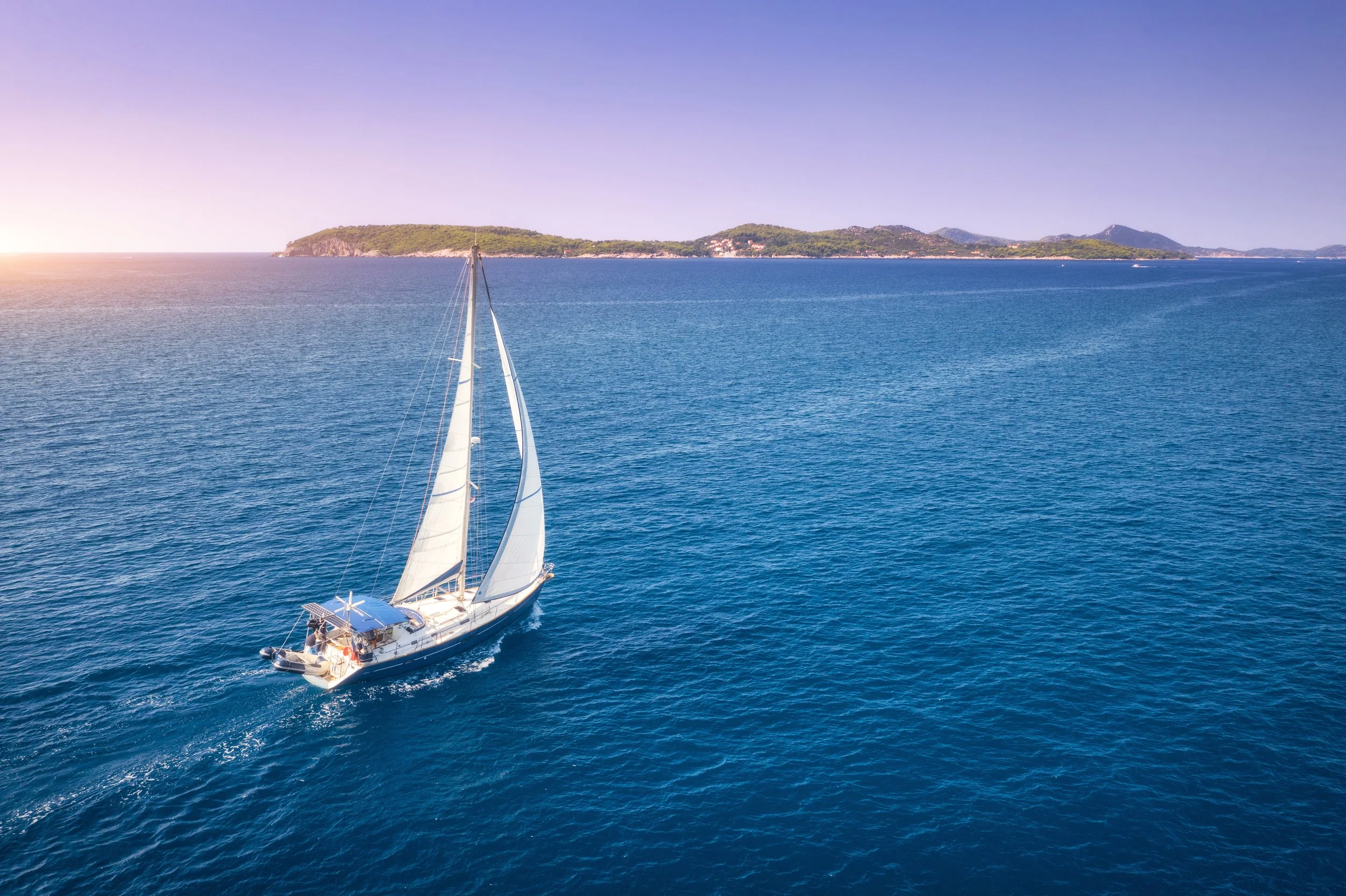 A sailboat sailing on calm blue ocean waters near a hilly, green island with a few buildings, under a clear sky at sunset.