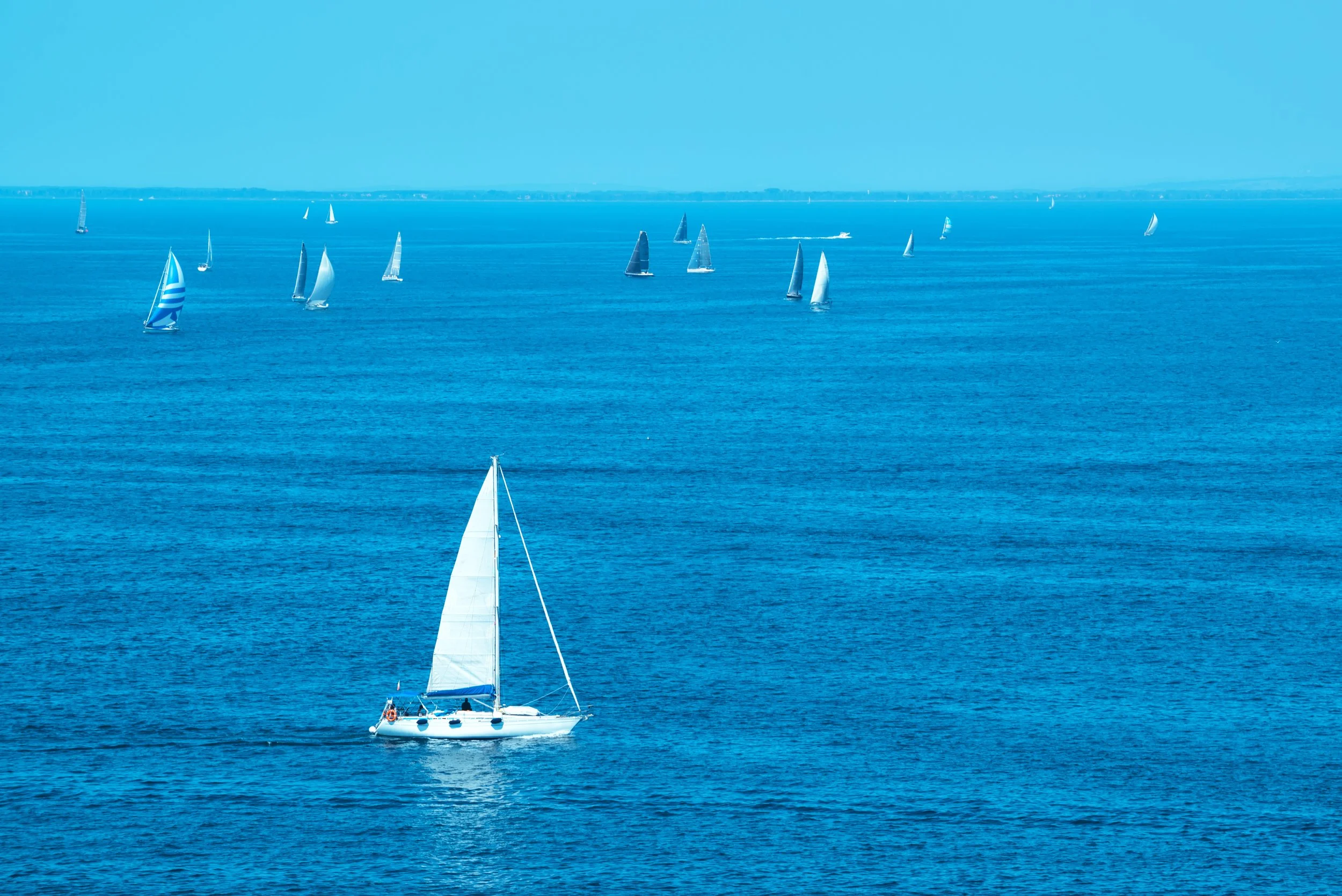 A sailboat with white sails on a calm blue ocean, with more sailboats in the distance and a clear sky.