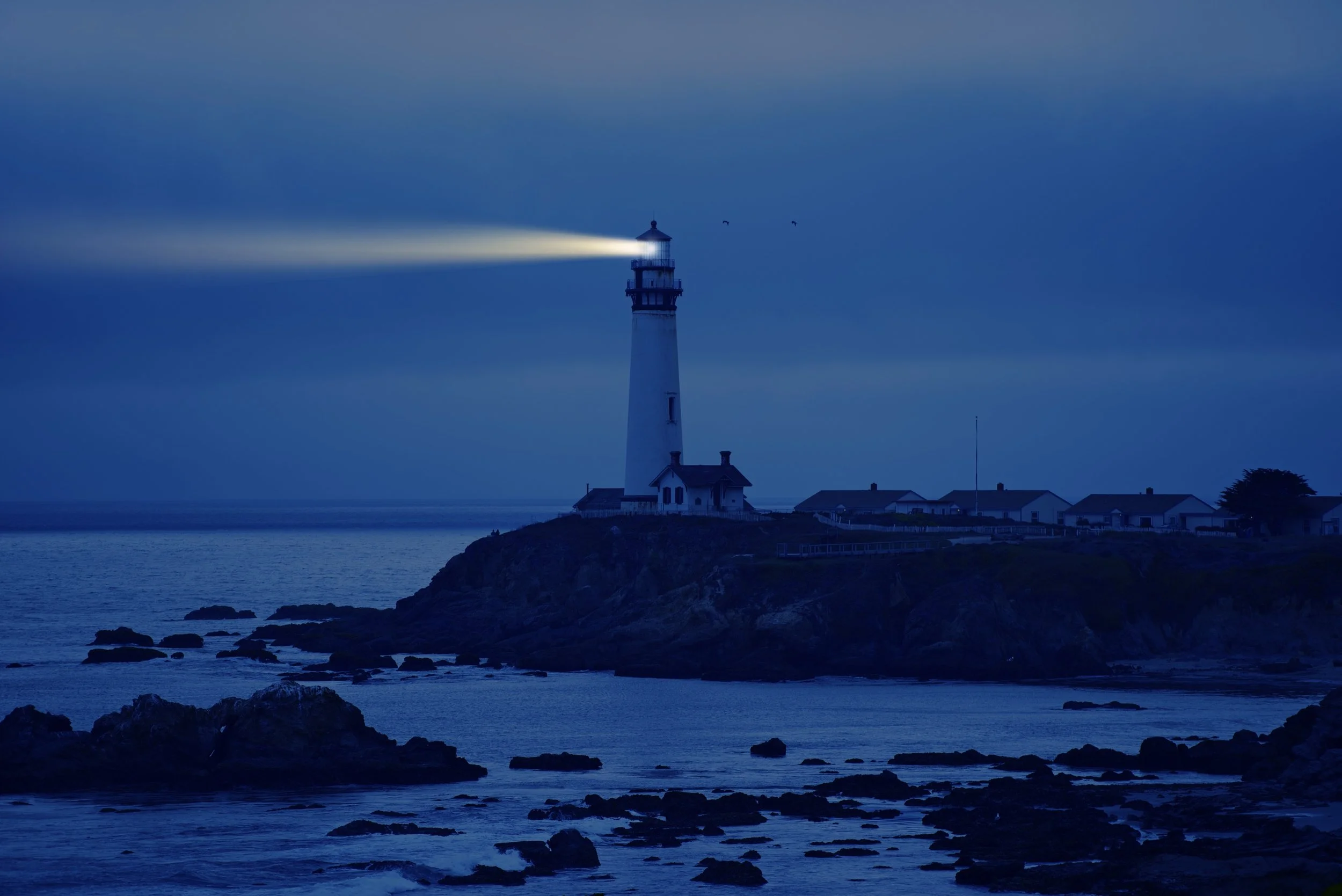 A lighthouse on a rocky coastline at dusk, with its light shining across the water. The sky is dark with a few clouds and two birds are flying nearby.