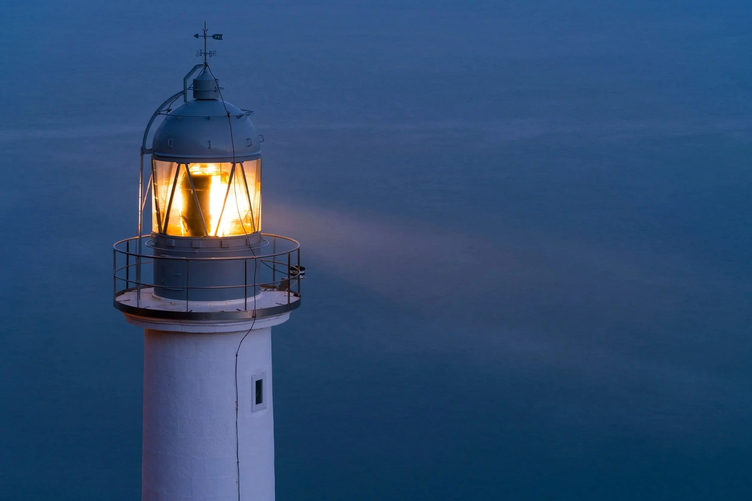 A lighthouse with its light on, located next to a body of water during dusk or dawn.