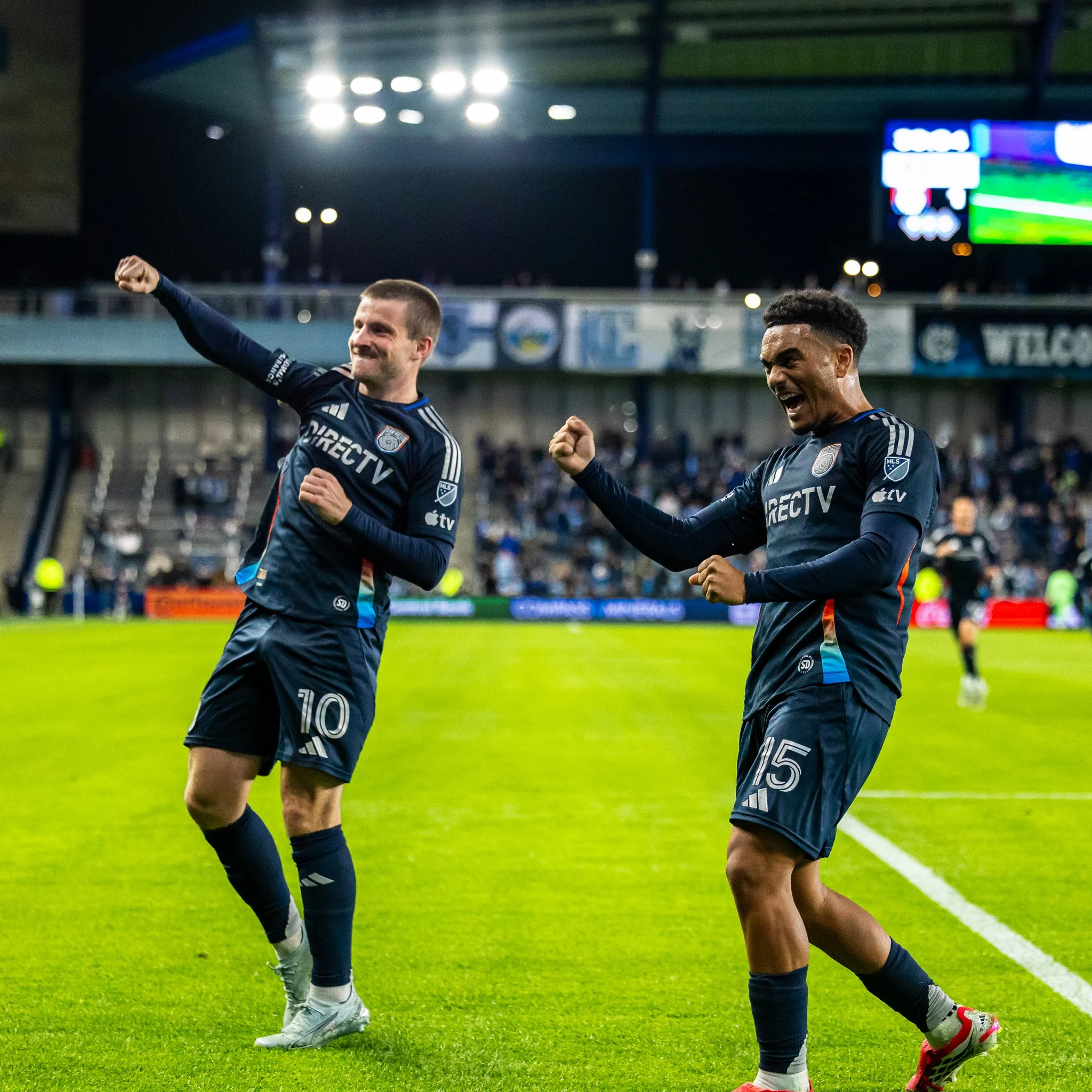 Two soccer players in dark blue uniforms celebrating on a soccer field at night, with stadium lights overhead and a digital scoreboard in the background.