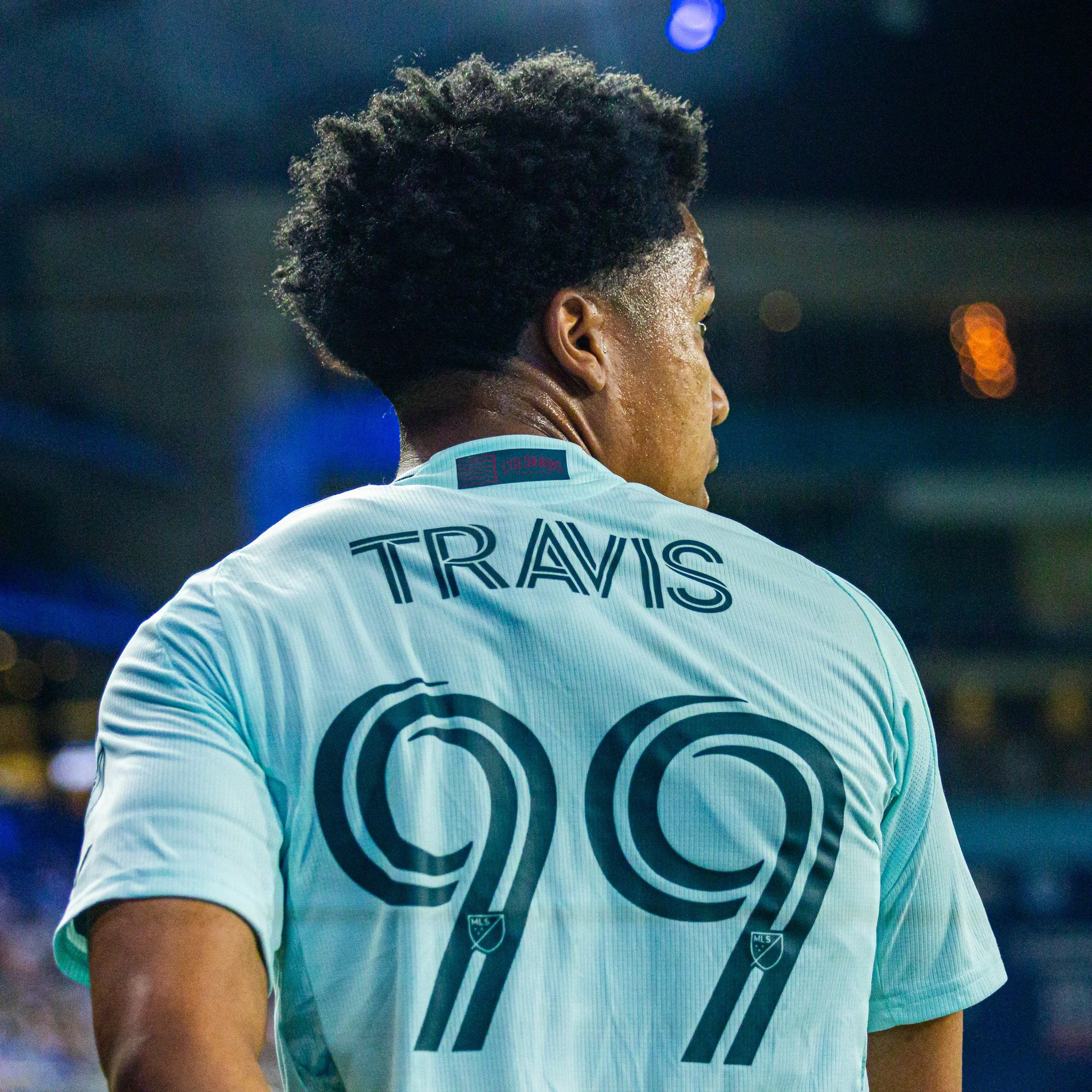 Close-up of a soccer player's upper back and shoulder, showing a white jersey with black and red accents, and black text reading 'Travel' and other words. The player has short curly black hair and is in a stadium or sports field at night.