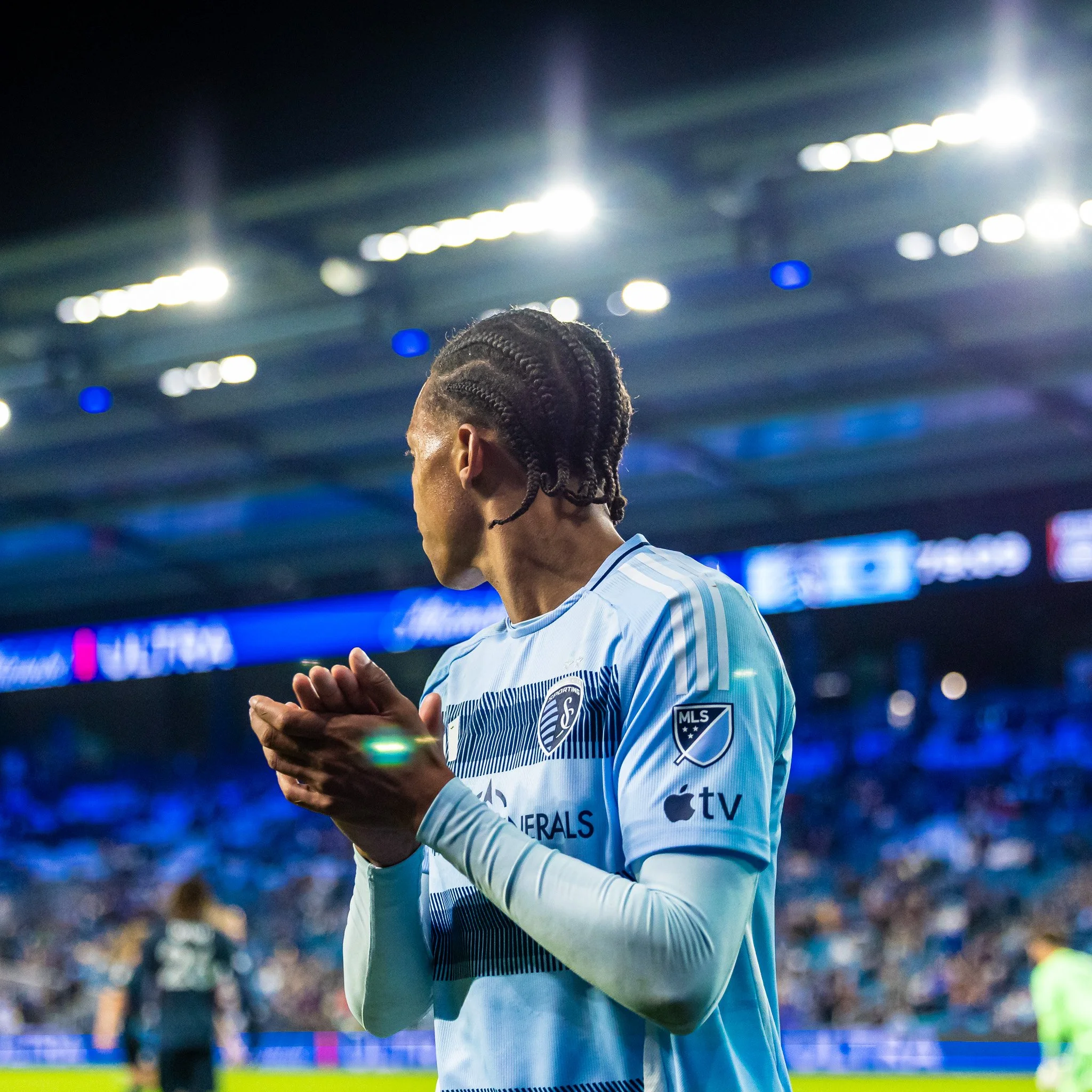 A soccer player wearing a light blue jersey with black and white details and braided hair, appears to be praying or concentrating during a match in a stadium.
