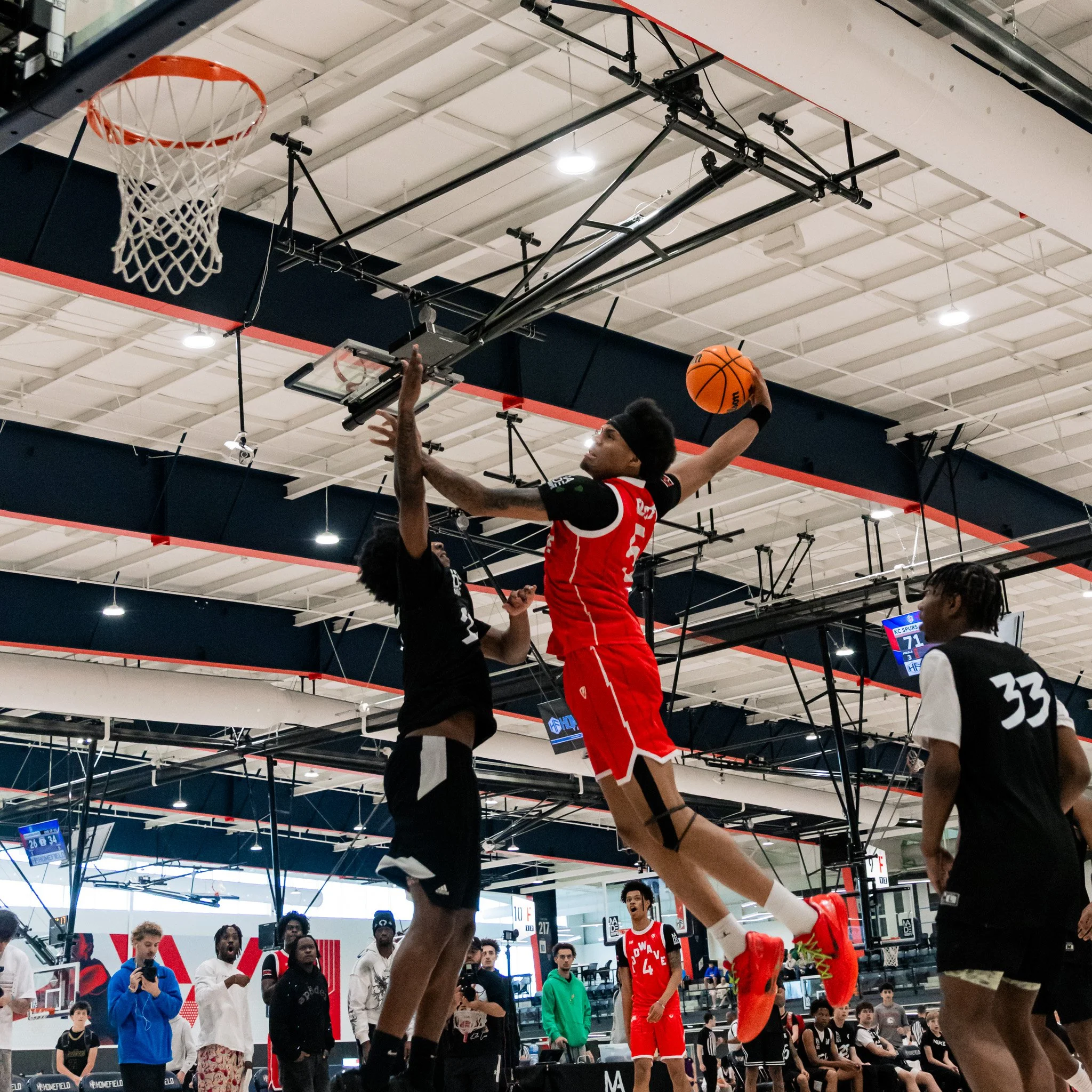 A basketball player in a red uniform is dunking the ball into the hoop during a game at an indoor gym. Several spectators and other players watch, with some taking photos or recording. The gym has high ceilings with bright lighting and digital scoreb