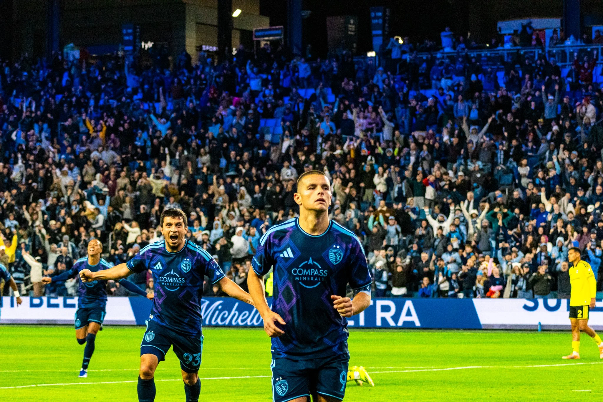 Soccer players in blue uniforms celebrating on the field during a nighttime match with a large crowd in the stands.