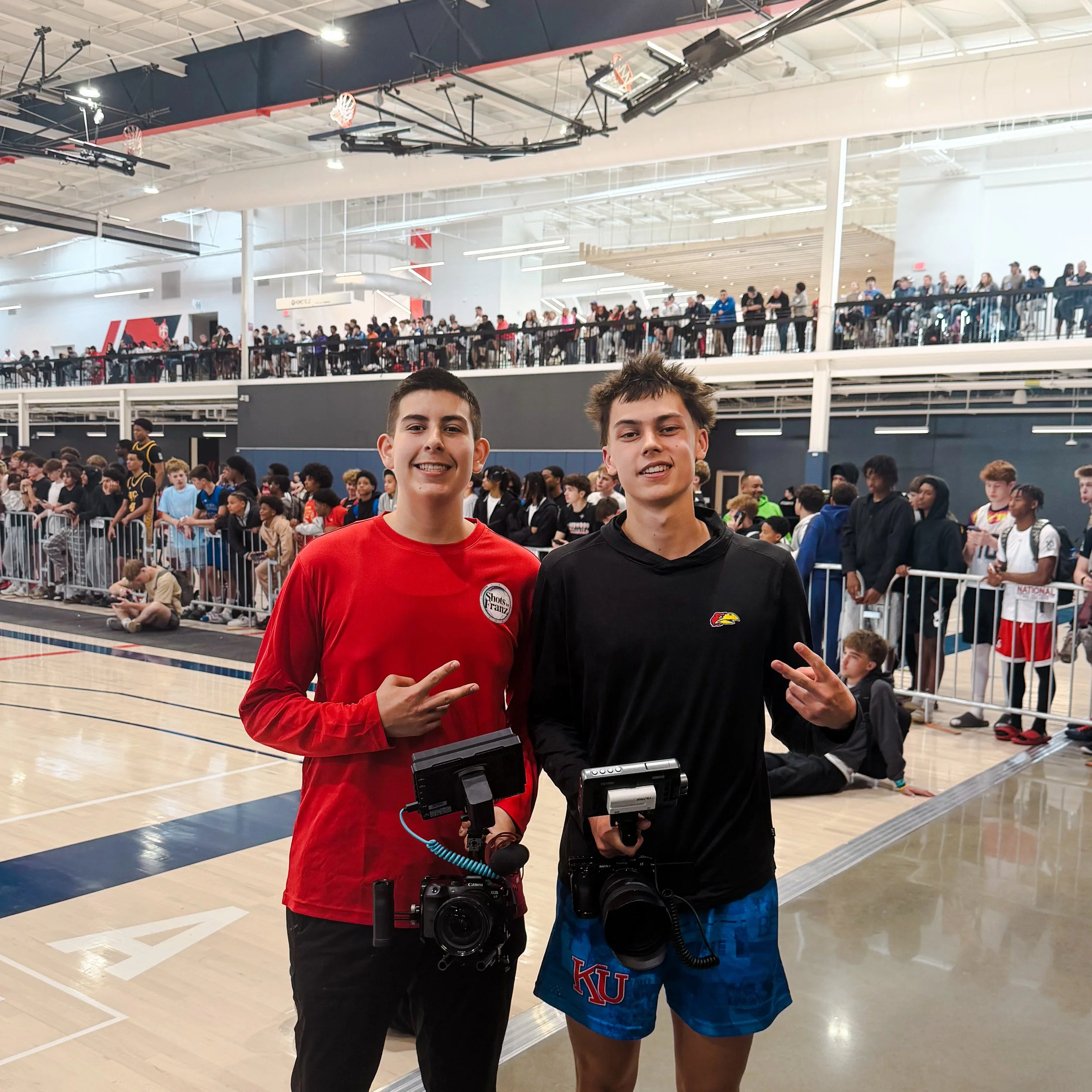 Two teenage boys standing in a crowded indoor sports arena, smiling, with spectators in the background. Each boy has a camera hanging from his neck, and they are making peace signs with their hands.