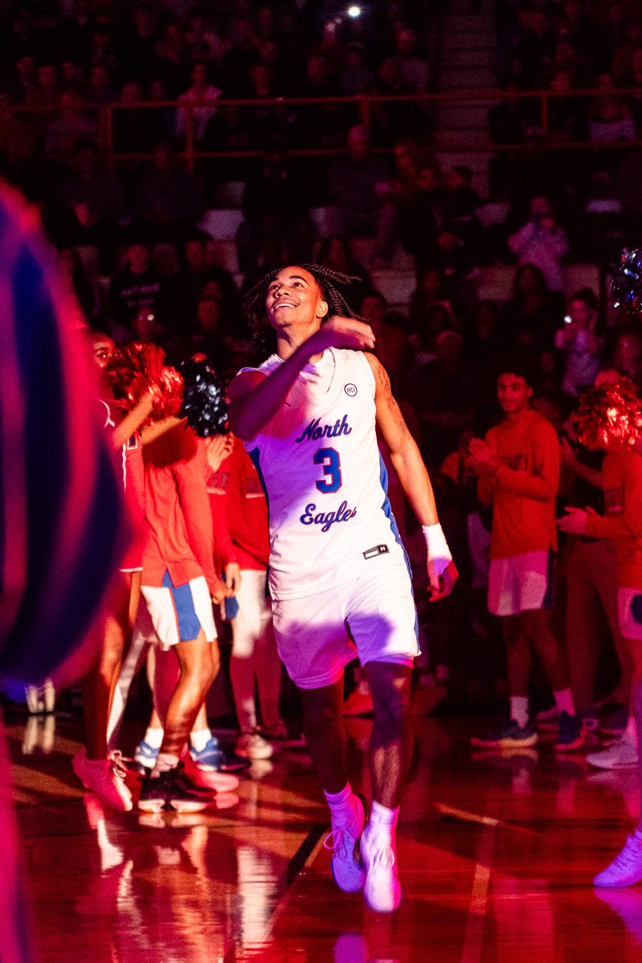 A basketball player from North Eagle wearing jersey number 3, celebrating on the court with fans and cheerleaders in the background.