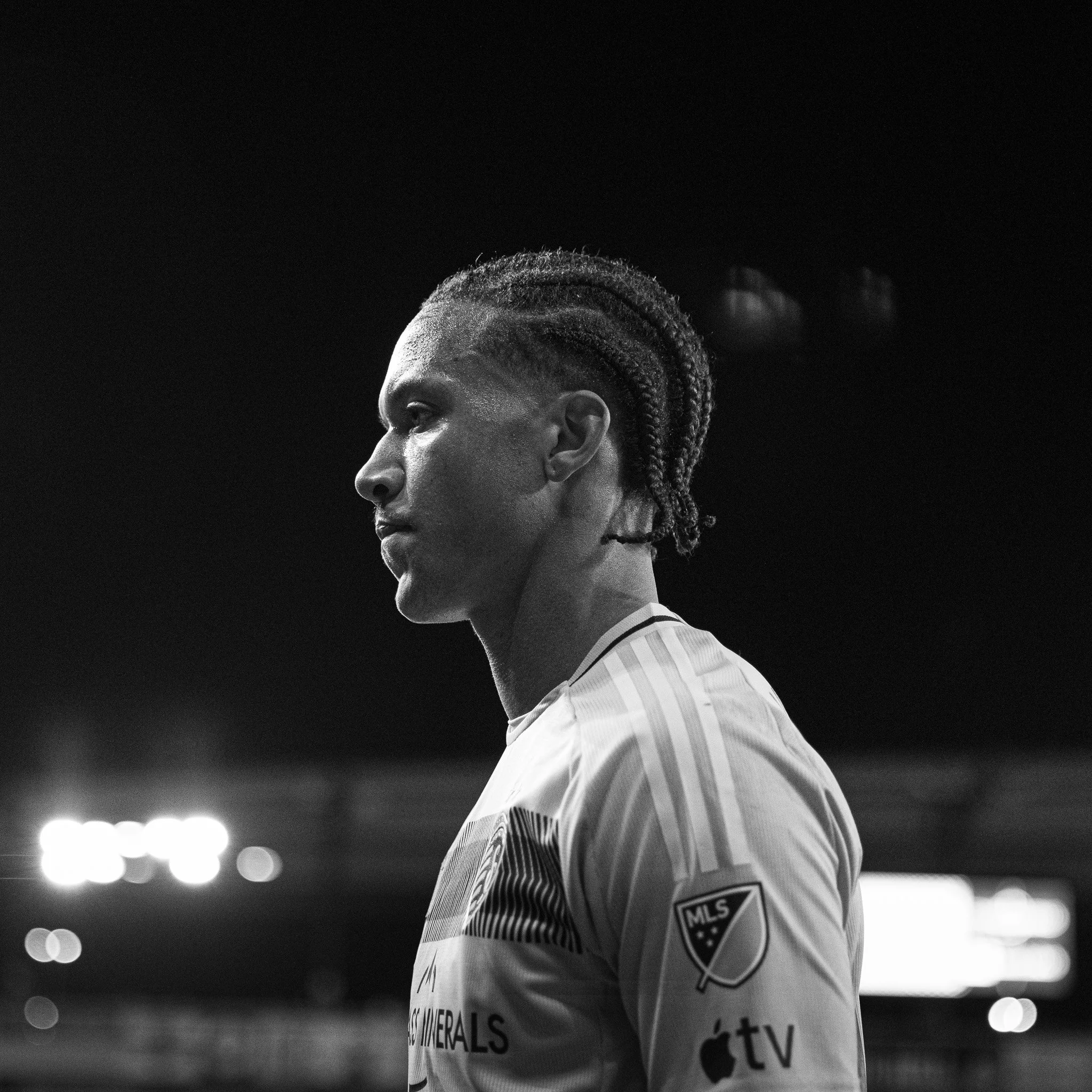 Close-up of a soccer player with braids on his head, wearing a white jersey with the MLS logo, standing on a field under stadium lights in black and white.