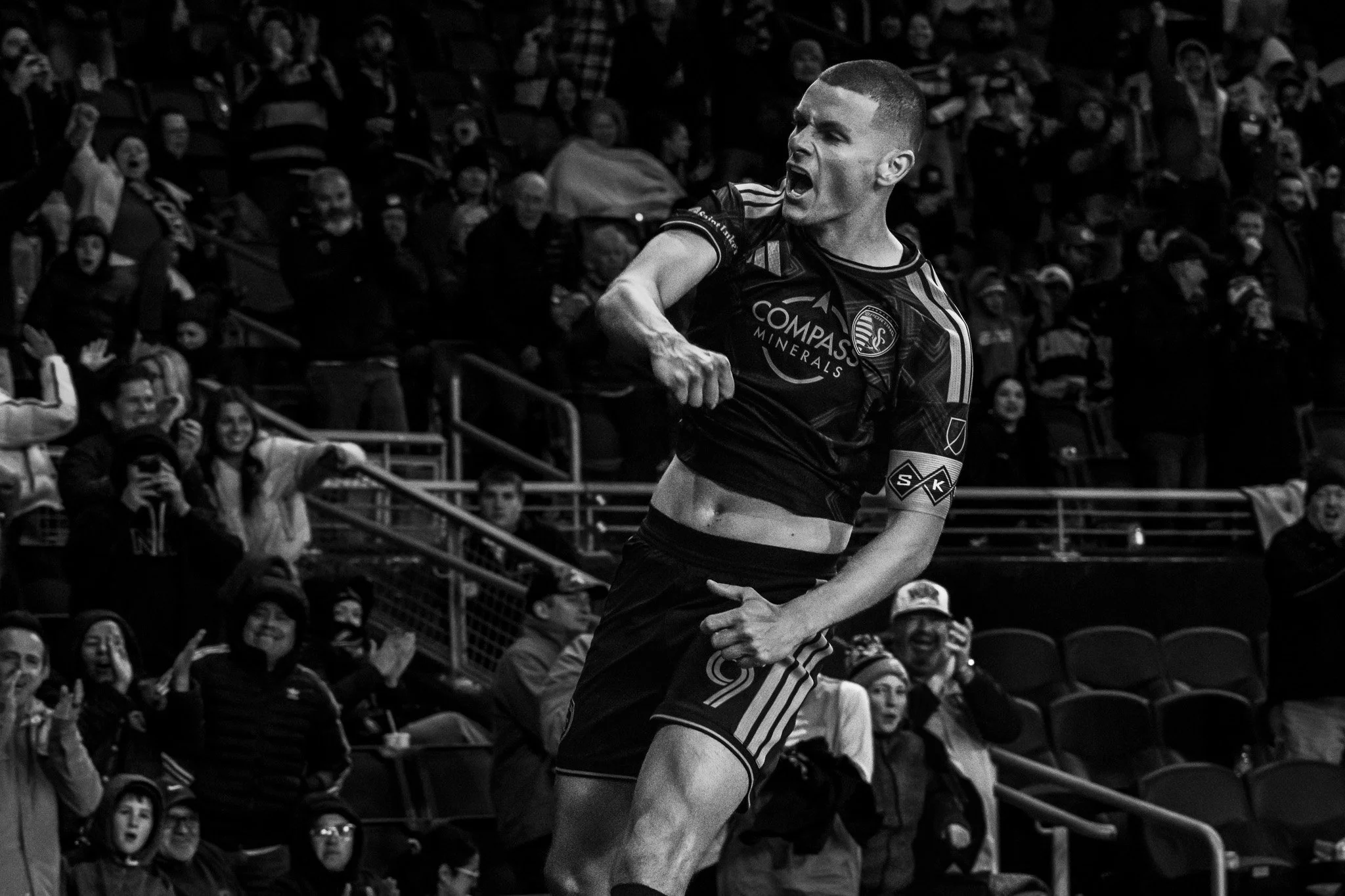 A male soccer player with short hair, wearing a dark jersey with logos, is celebrating on the field with his mouth open and one arm extended to the side. The stands behind him are filled with cheering spectators.