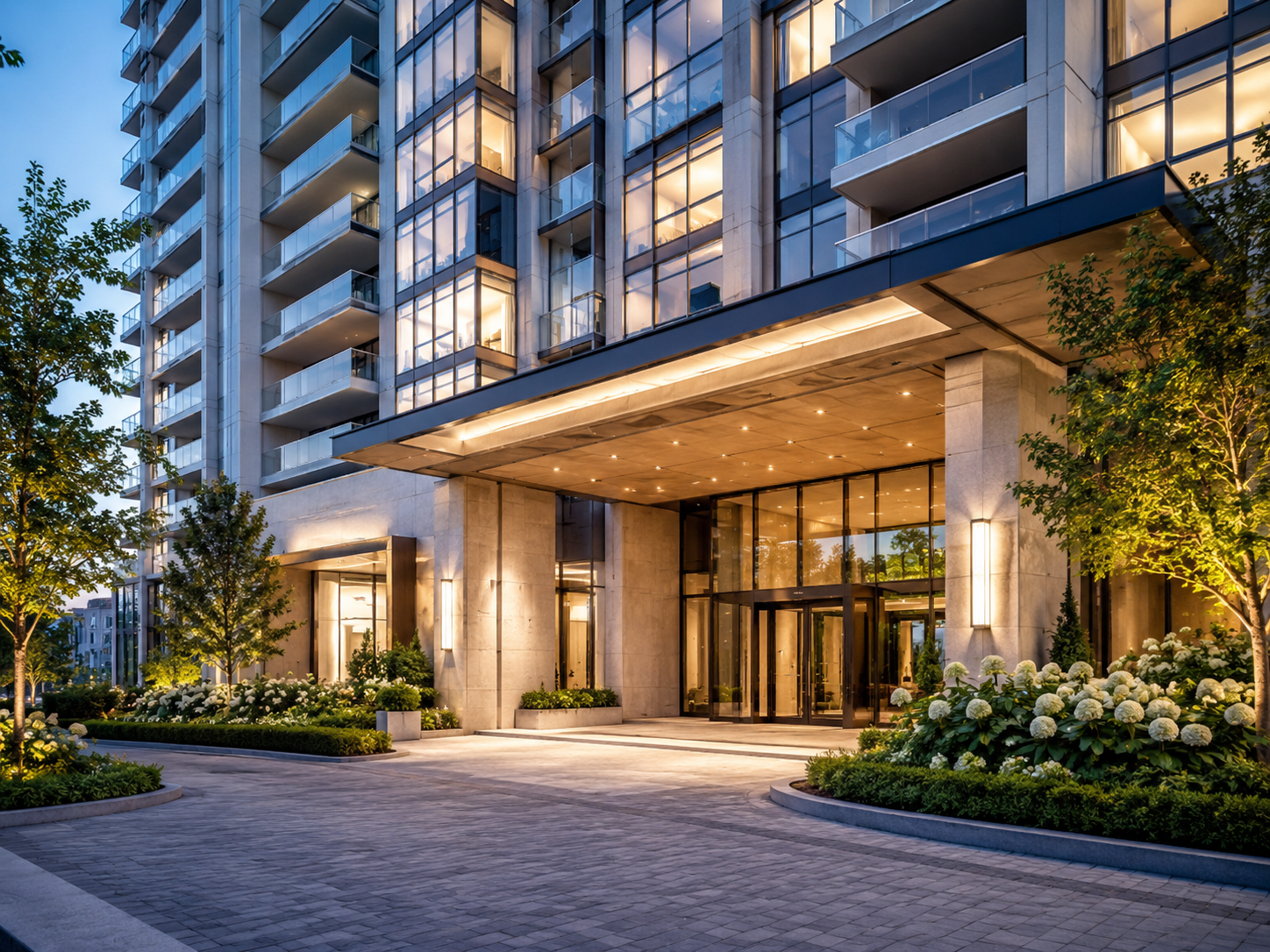 Exterior view of a modern high-rise building with glass windows and balconies, illuminated at dusk, with landscaped greenery and flowering plants at the entrance.