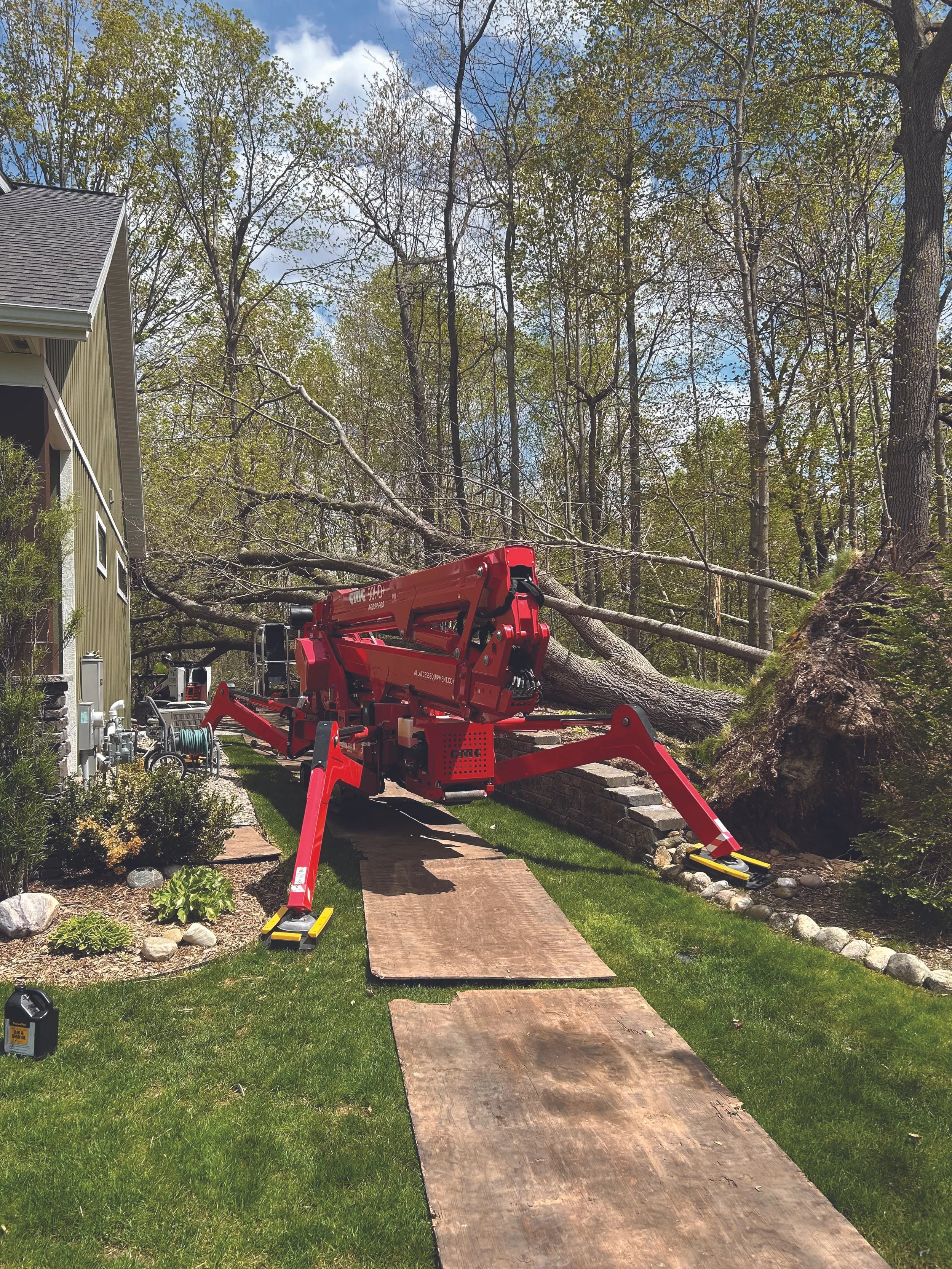 A fallen tree on a residential property with a red lift equipment nearby, a brick pathway, green lawn, and a house on the left, under a partly cloudy sky.