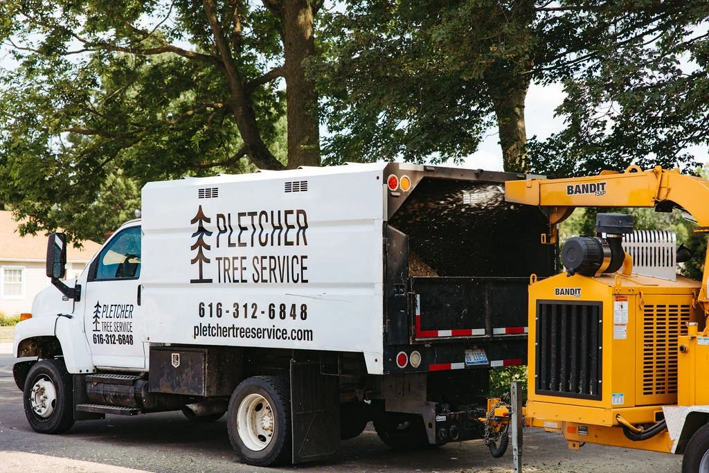 A white tree service truck with the company name Plexcher Tree Service, phone number, and website. A yellow wood chipper attached to the truck, parked on a residential street with trees in the background.