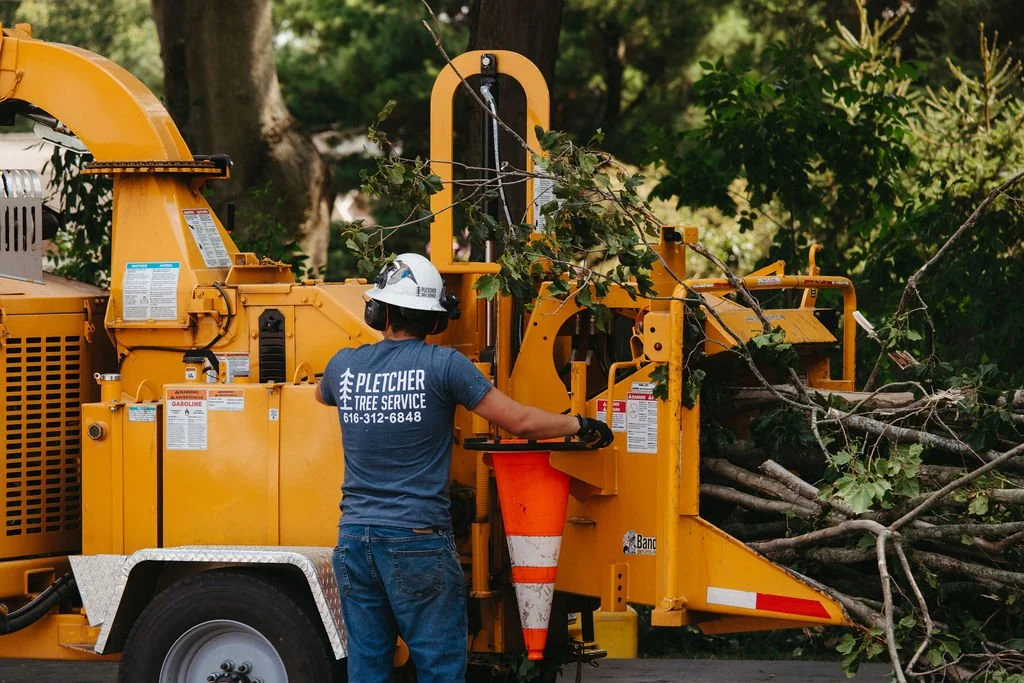 Worker operating a tree chipper, processing branches and logs, on a street with lush greenery in the background.