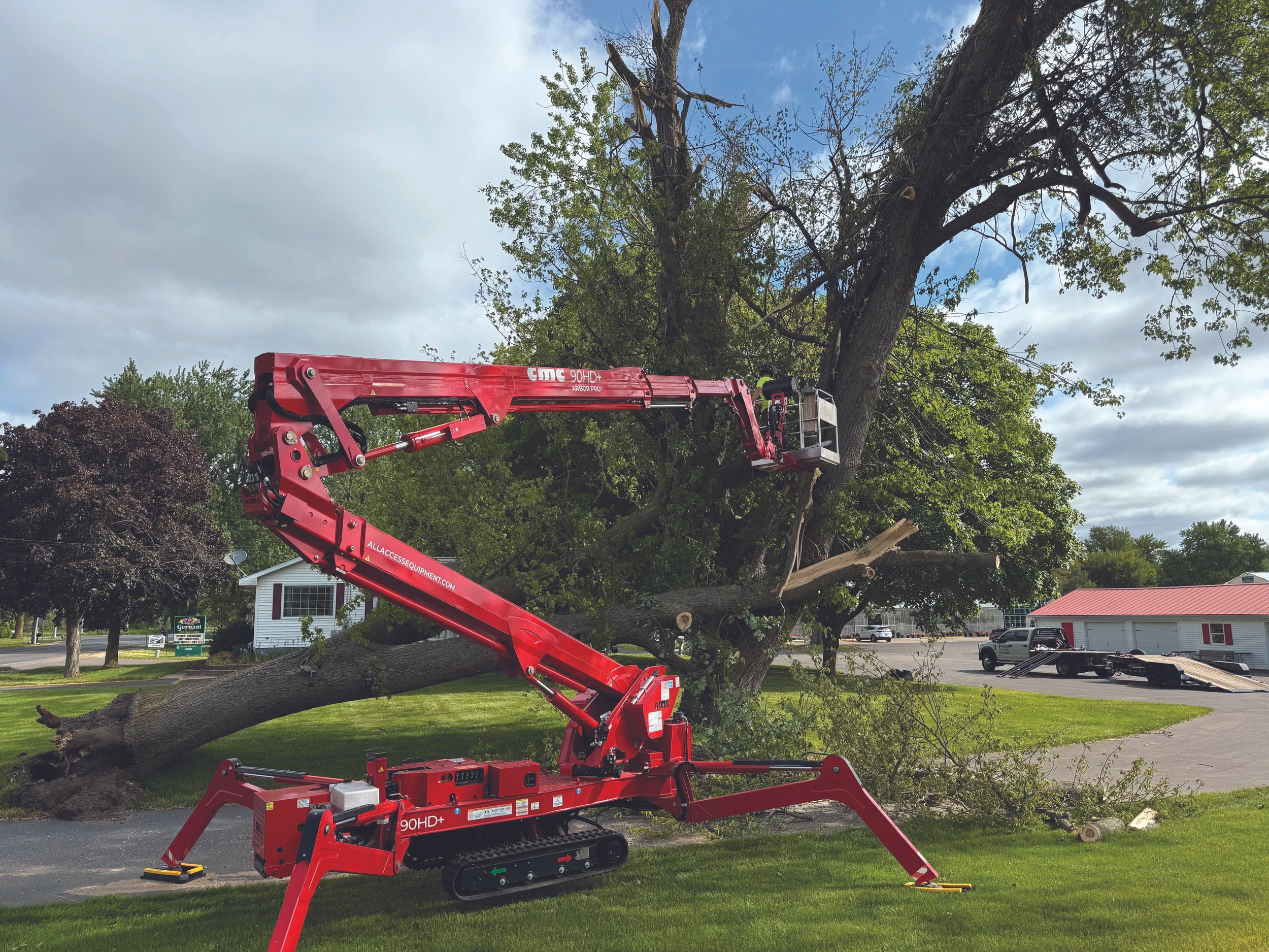 A red hydraulic lift with a bucket platform raised to trim a large tree in a suburban neighborhood. The tree has green and some broken branches. There are houses and parked cars in the background, and the sky is partly cloudy.
