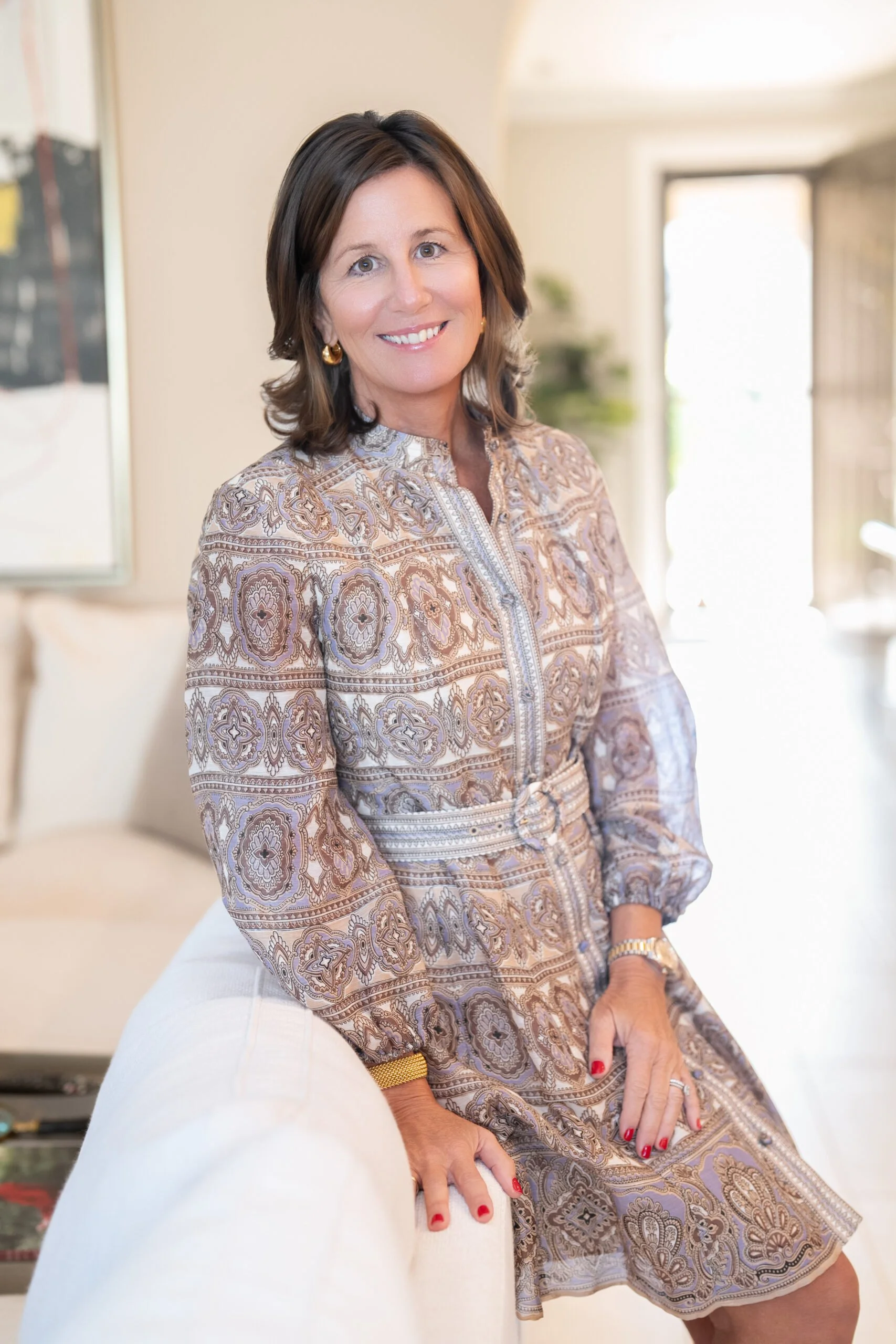 A photo of Lisa Baird Hoey, relationship coach; a woman with shoulder-length dark brown hair, wearing gold earrings and a paisley patterned dress, smiling while sitting on a white chair in a bright room with a blurred background.