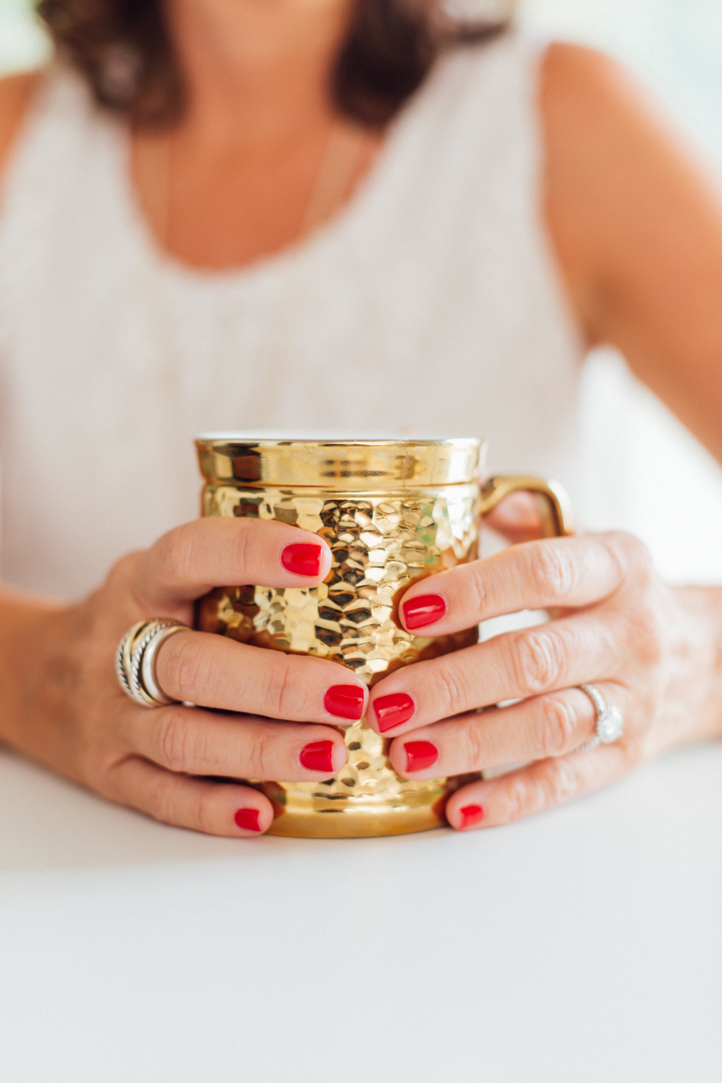 Person with red painted nails holding a gold textured mug with both hands, wearing rings, sitting at a white surface with a blurred background.