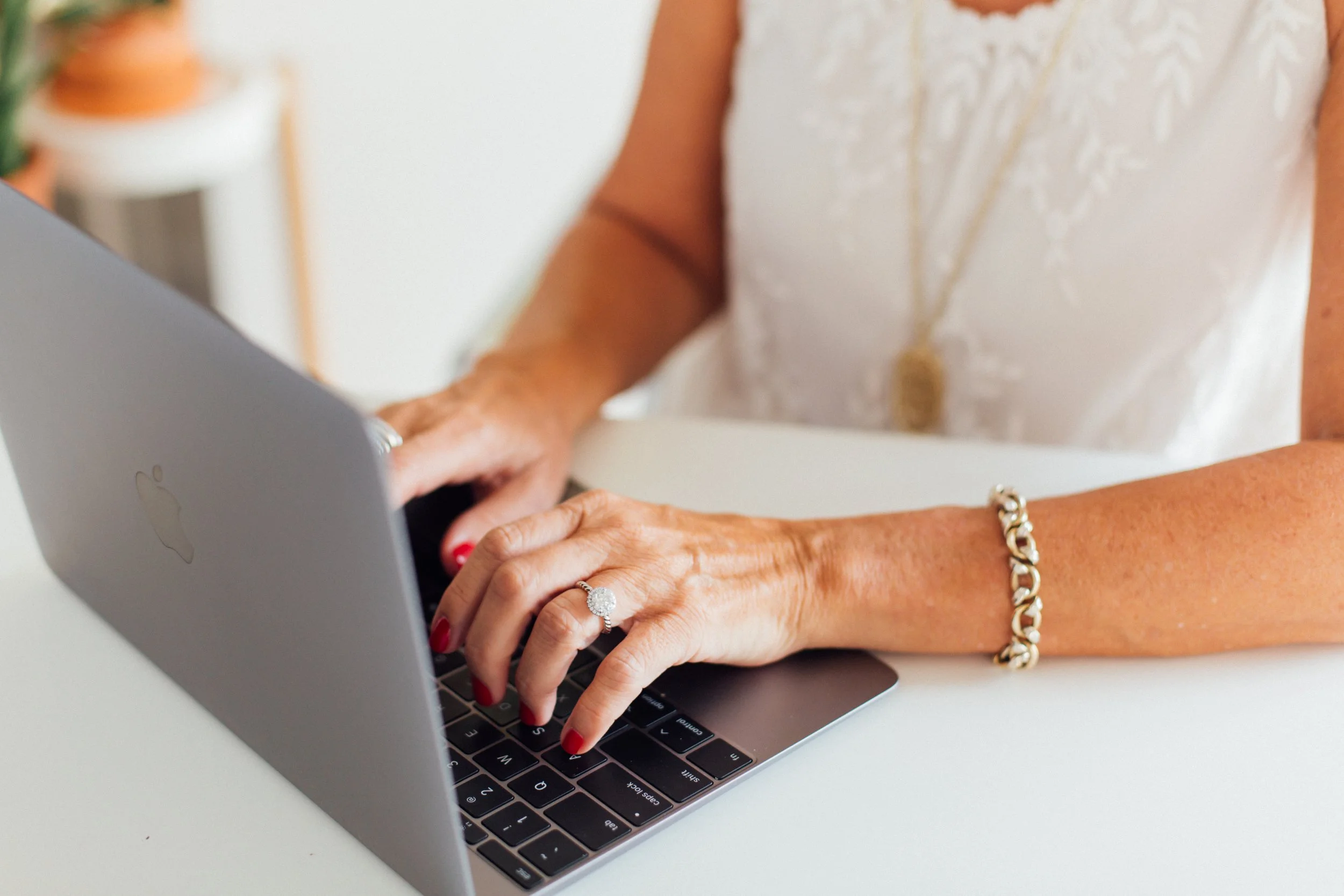 Close-up of a person's hands typing on a MacBook laptop keyboard. The person is wearing a gold bracelet and a ring, and has red nail polish. The background shows part of a blurred decorative object.