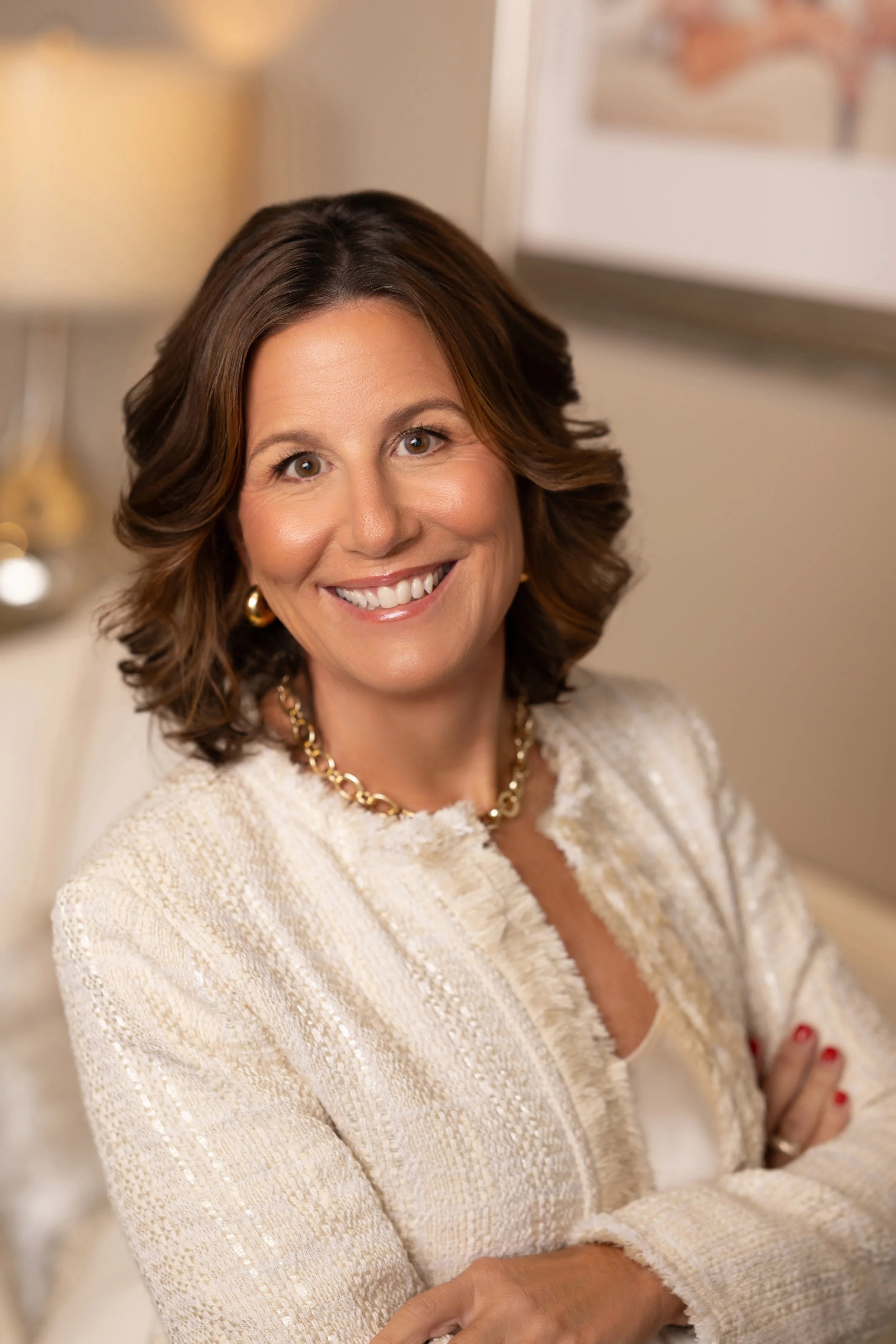 A smiling woman with shoulder-length brown hair wearing a cream-colored, textured jacket and gold jewelry.