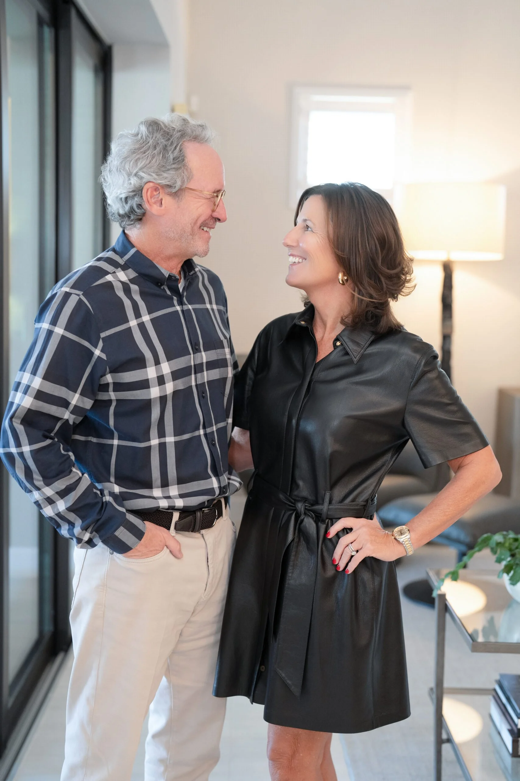 A man with gray hair, glasses, wearing a dark plaid shirt and beige trousers, smiling and standing close to a woman with shoulder-length brown hair, wearing a black leather dress, smiling back, in a brightly lit modern room with a window and a lamp in the background.