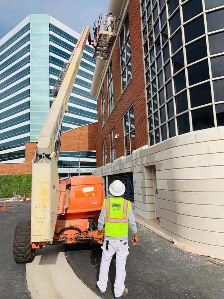 Construction worker in a white hard hat and neon yellow safety vest standing next to an orange boom lift, working on the exterior of a modern multi-story building with brick and glass windows.