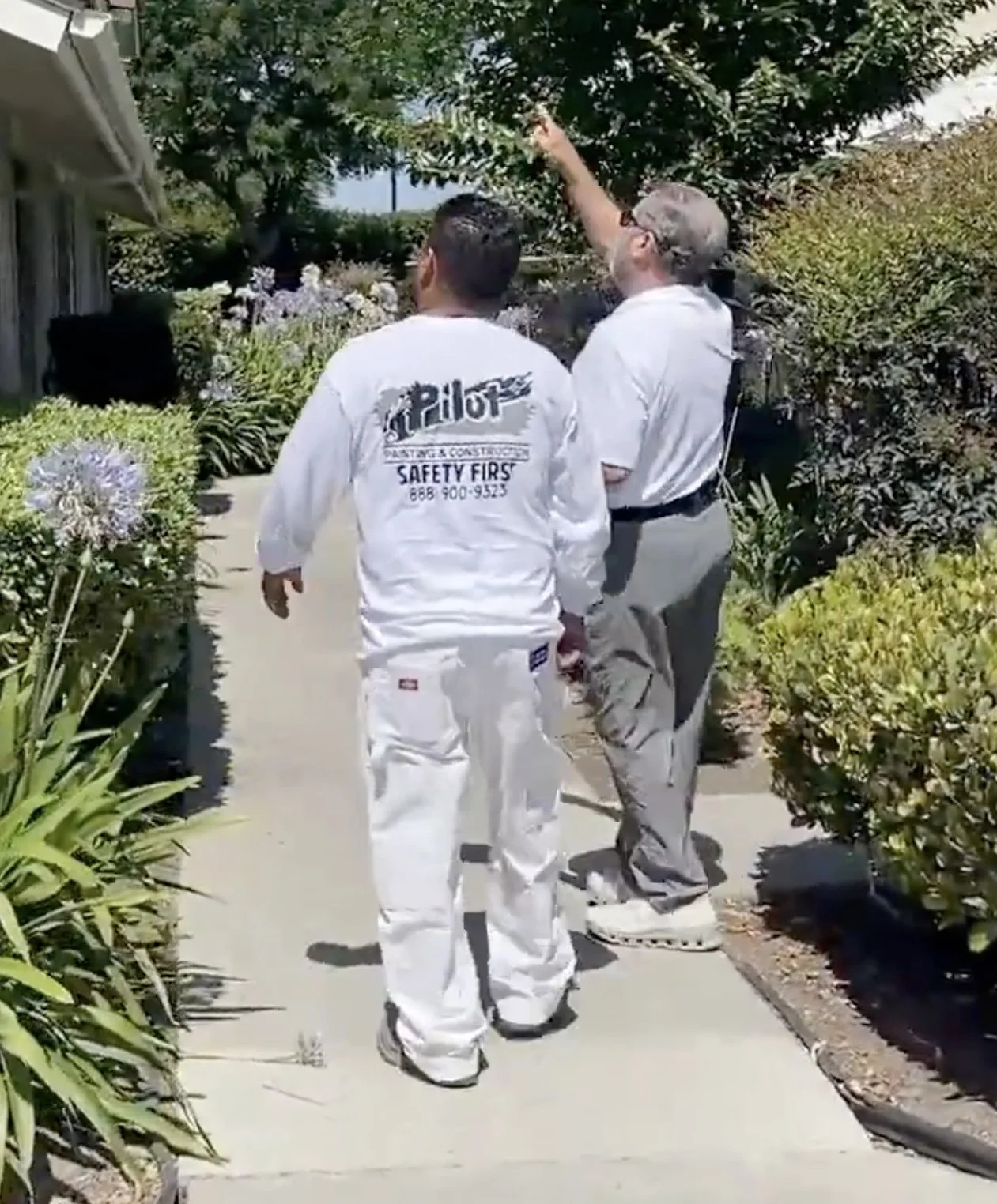 Two men walking on a sidewalk among landscaped bushes and flowers; one is wearing a white uniform with 'Painter & Contractor' and 'Safety First' written on the back, and the other is wearing a white shirt and light gray pants, pointing to a tree.