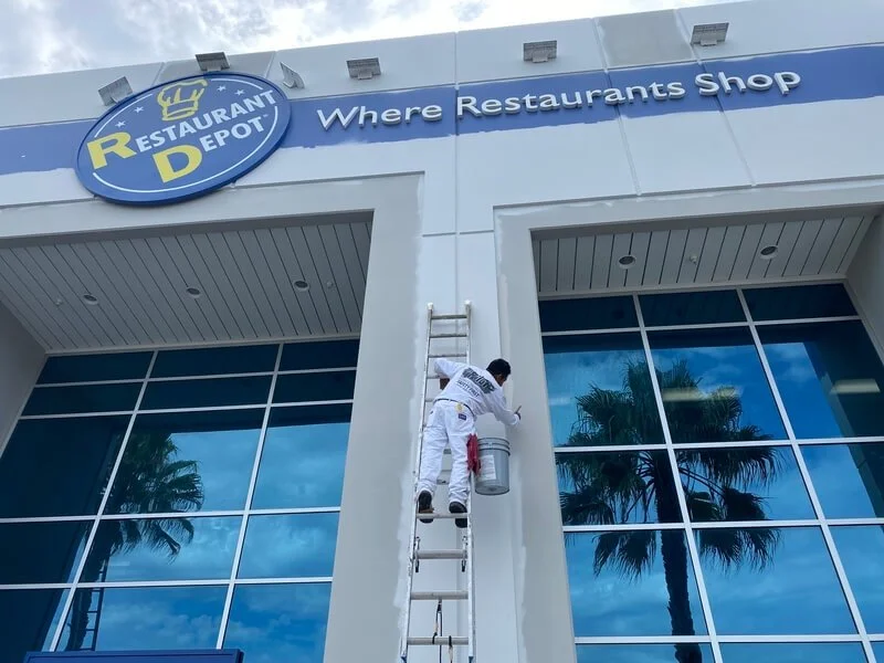 Worker painting the exterior of a Restaurant Depot store, with a large sign that reads 'Where Restaurants Shop' and windows reflecting palm trees.