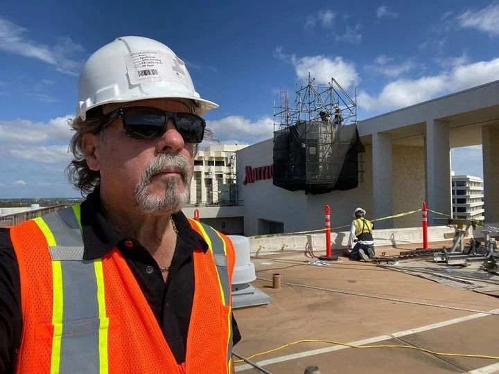 Construction worker wearing sunglasses, a white safety helmet, and an orange safety vest on a building rooftop under a blue sky with scattered clouds, with construction equipment and workers in the background.