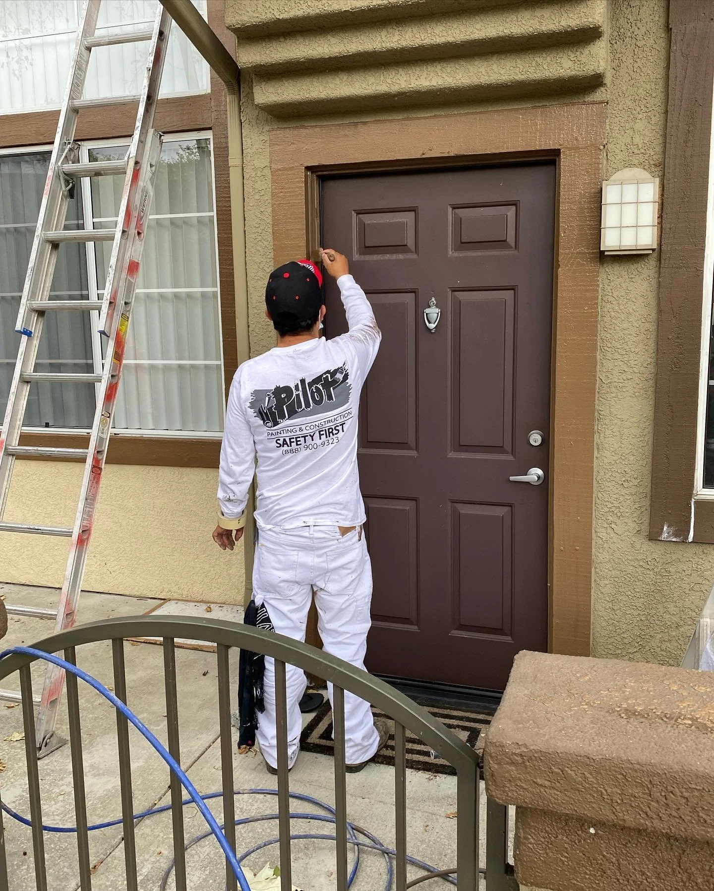 A worker painting a brown door outside a building. The worker is wearing white pants and a white shirt with text on the back. There are ladders and a railing nearby, and a window with white curtains on the building.