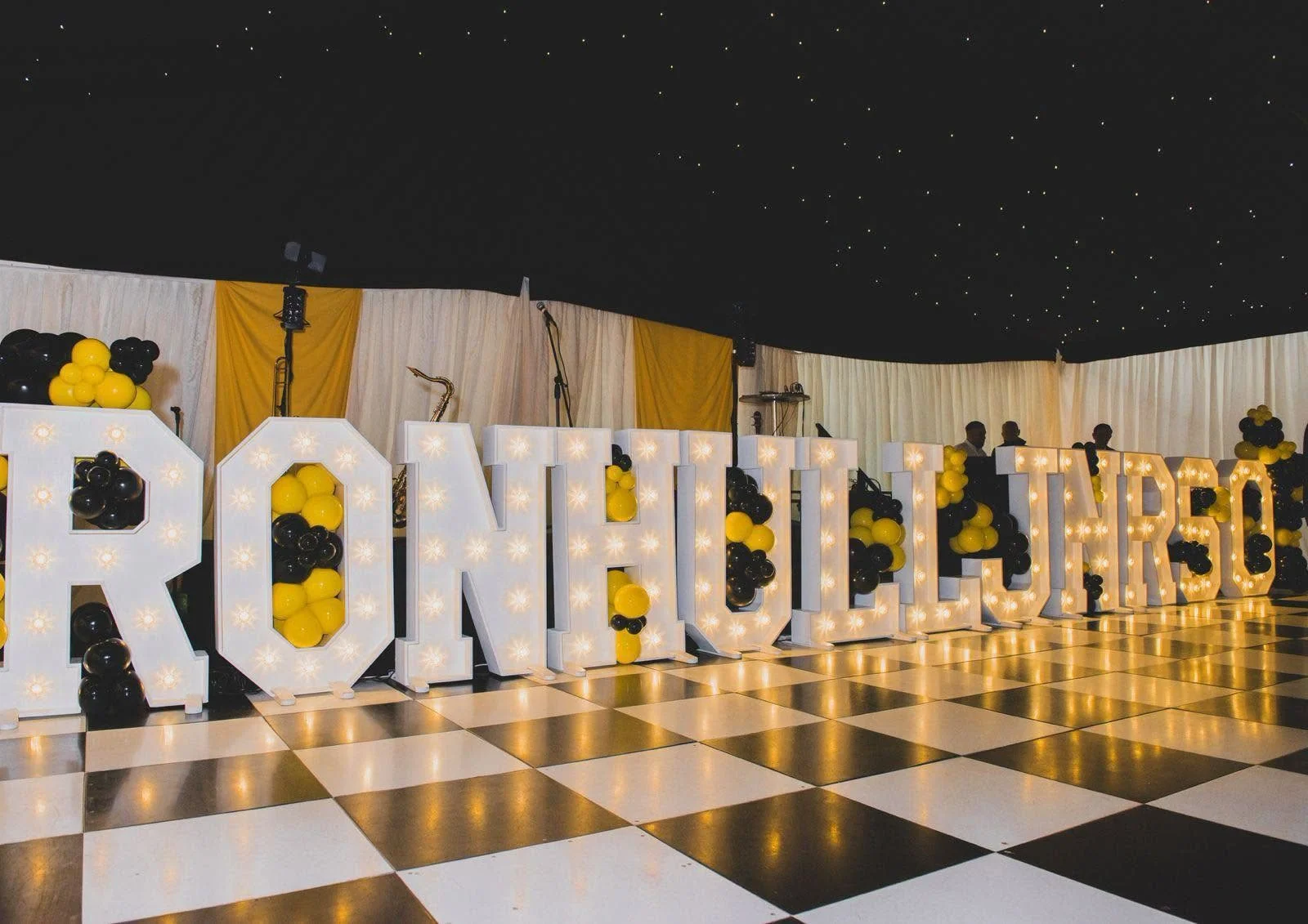Light-up RON HULL JNR 50 letters with yellow and black balloons beside a black and white dance floor
