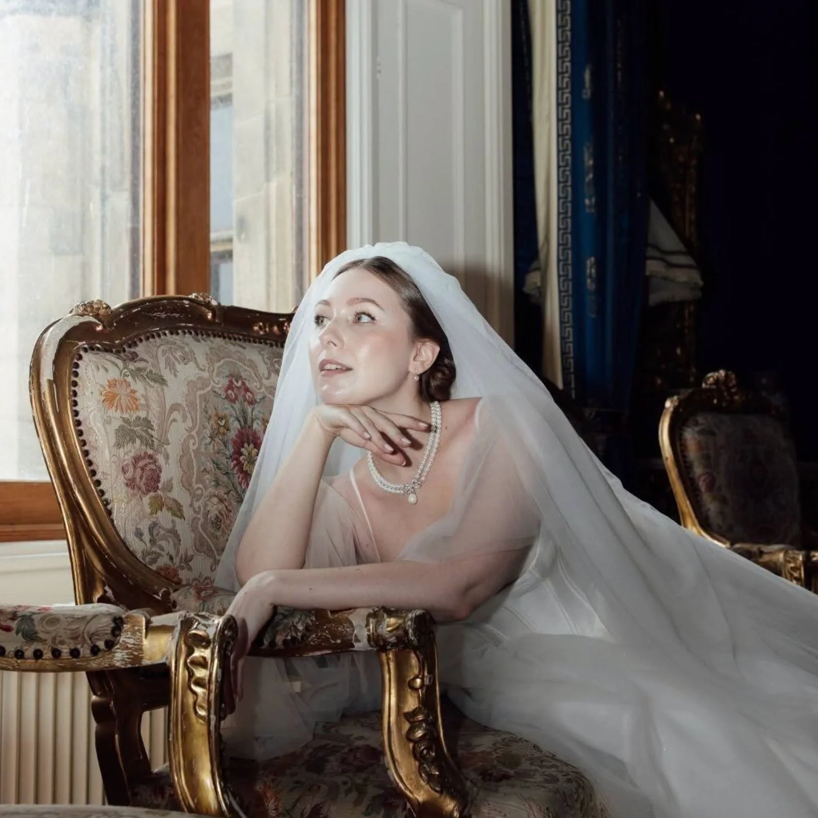 Bride in a veil seated on a gilt chair by a window