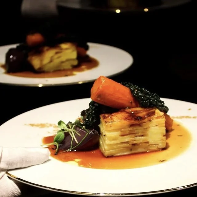 A plated gourmet dish with layered potato gratin, topped with a slice of carrot, accompanied by microgreens and dark sauce, on a white plate.