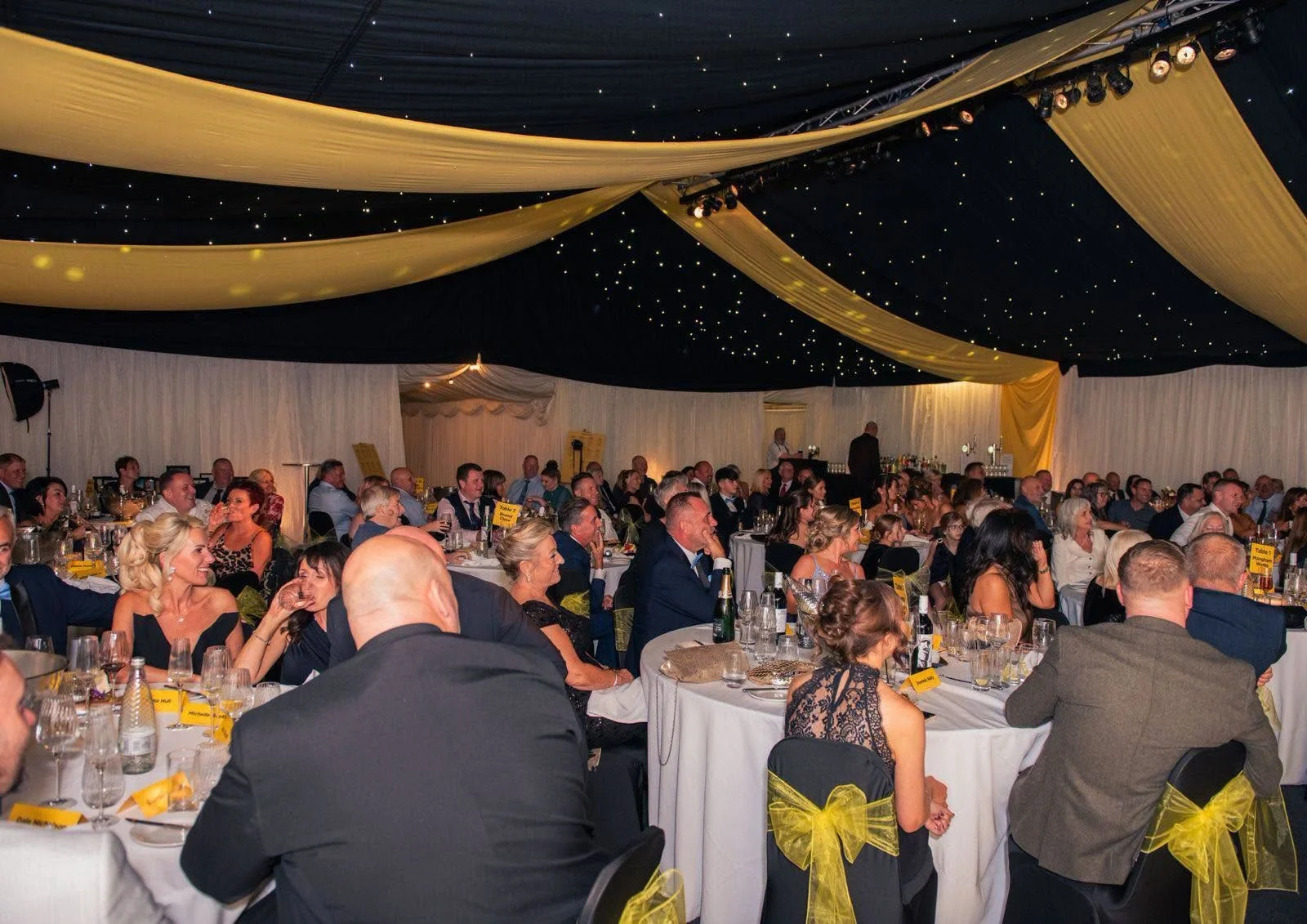 Guests seated at round tables in a starlit marquee with yellow drapes for the Ron Hull Jnr Ltd 50th anniversary dinner