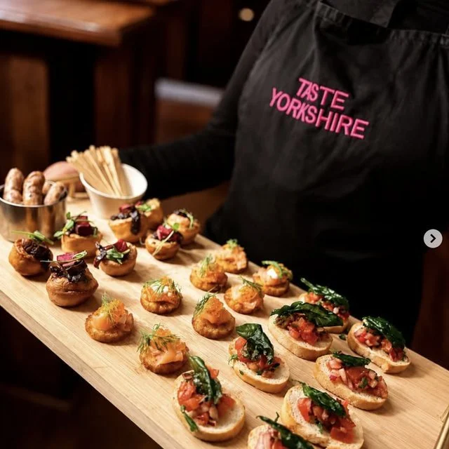 A person wearing a black apron with pink text reading 'Taste Yorkshire' presents a wooden serving board with an assortment of small appetizer bites, including topped crostinis and other garnished finger foods.