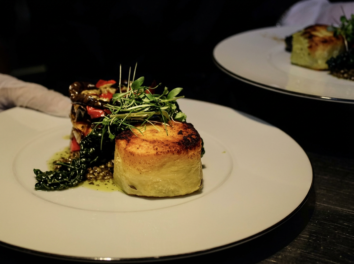 A gourmet plated dish featuring a seared scallop with a golden-brown crust, garnished with microgreens, accompanied by a vegetable medley of cooked greens and red peppers, and a sauce underneath.