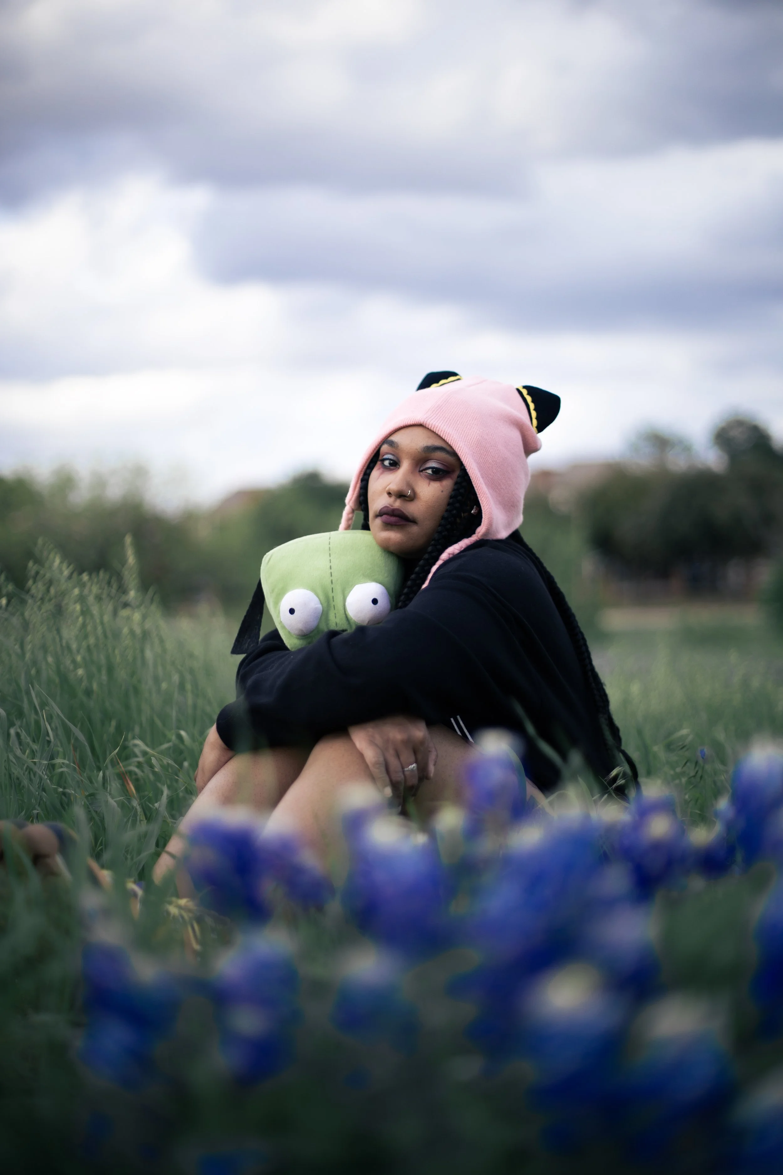 Local Dallas, Texas, DFW Non-traditional Fashion Editorial Creative Portraits Cover Art Photographer Photoshoot. Musician igimeji . A young woman with braided hair, wearing a pink knit hat with black ears, sits in a grassy field holding a green plush