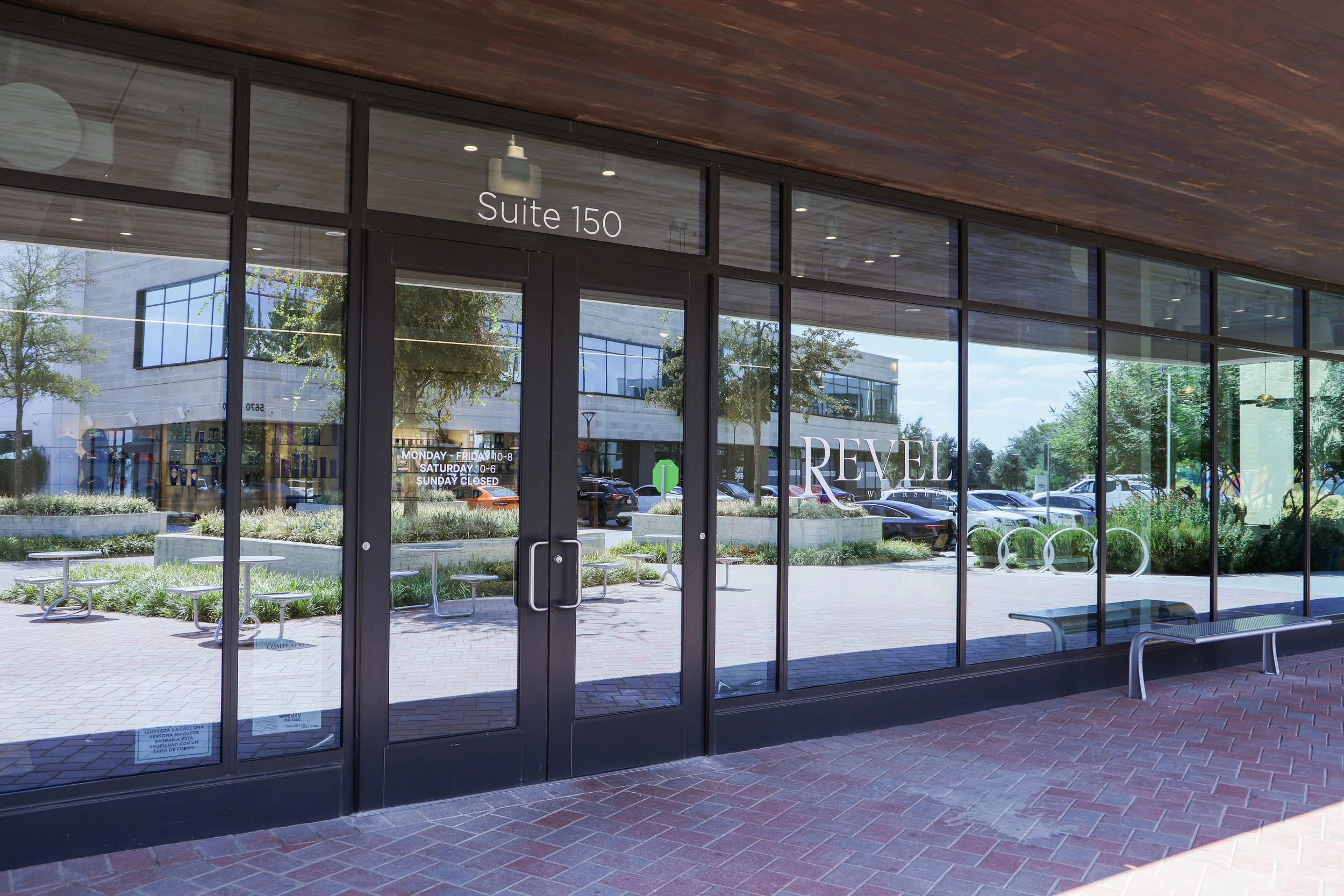The image shows a modern building entrance with large glass windows and doors marked 'Suite 150'. Outside, there are benches, small tables, and landscaped greenery along a paved area, reflecting trees and the building across the parking lot. The sky 