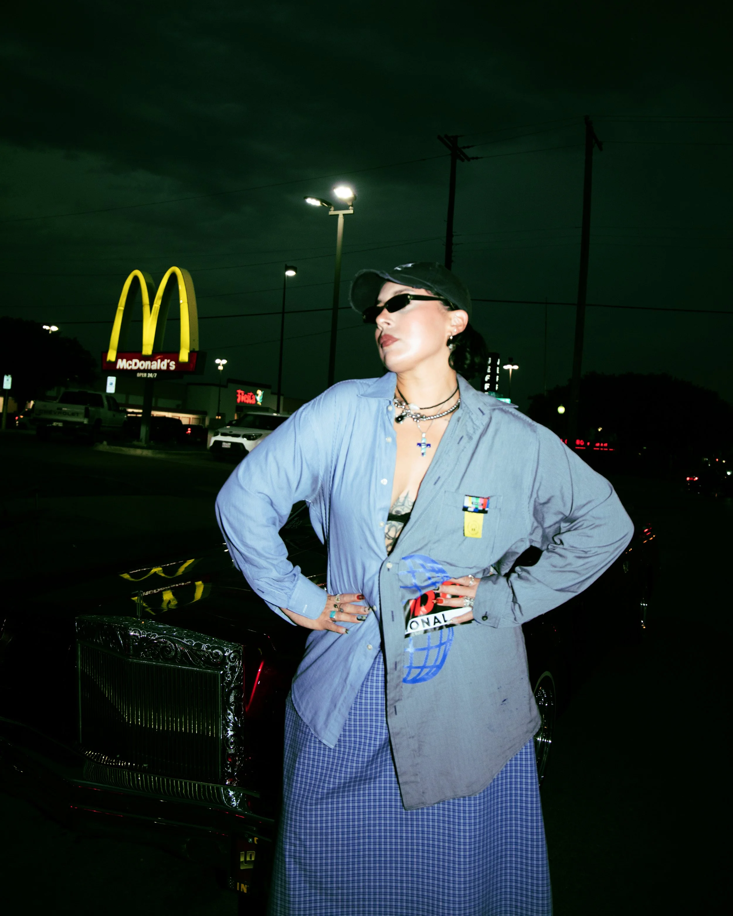 Local Dallas, Texas, DFW Non-traditional Fashion Editorial Creative Portraits Cover Art Photographer Photoshoot. Nataly Medina Crecer Dallas. Woman with dark hair, wearing sunglasses and a black cap, standing outdoors at night near a classic black ca
