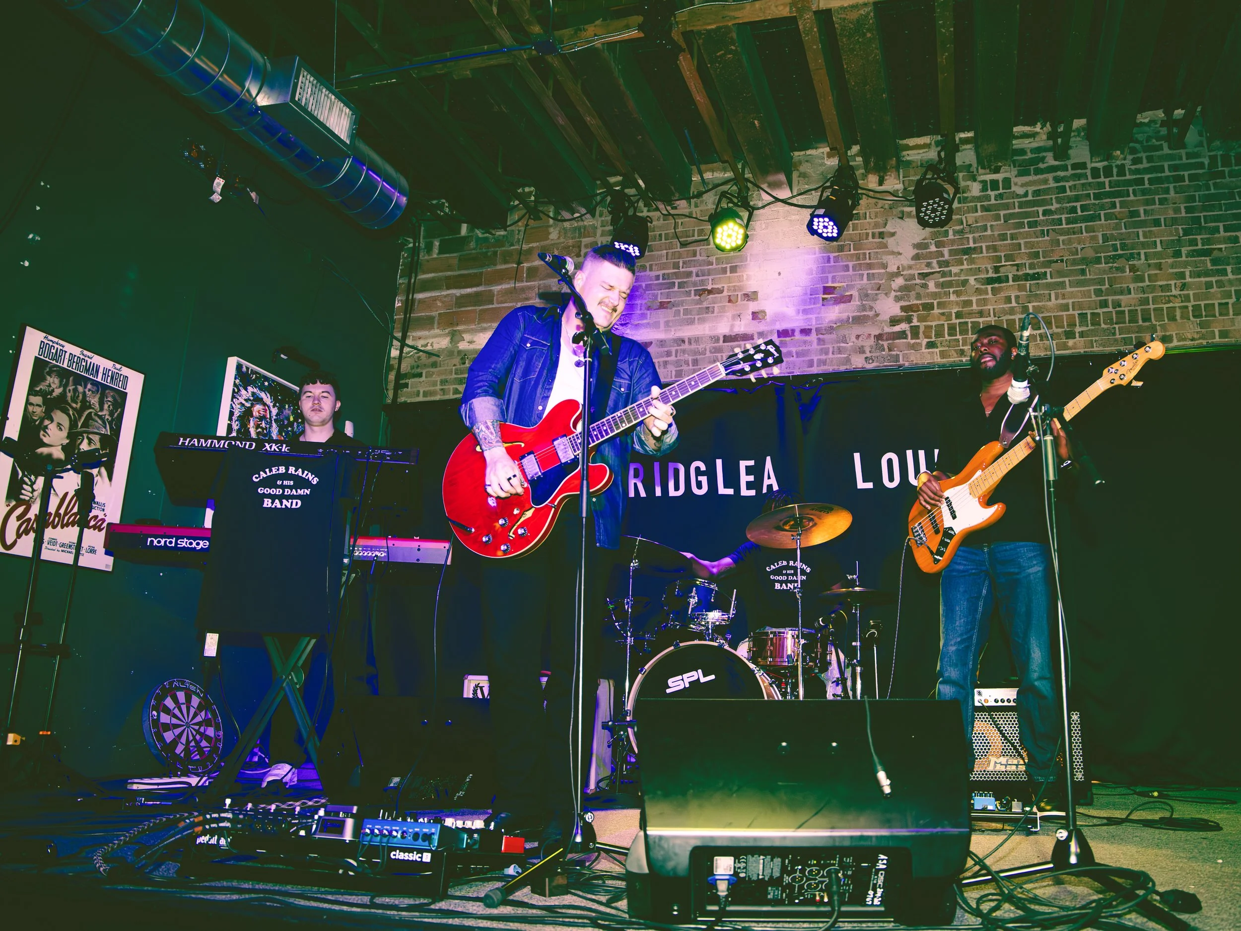 A band performs on stage in a dimly lit venue with a brick wall background, featuring a guitarist, bass player, keyboardist, and drummer.