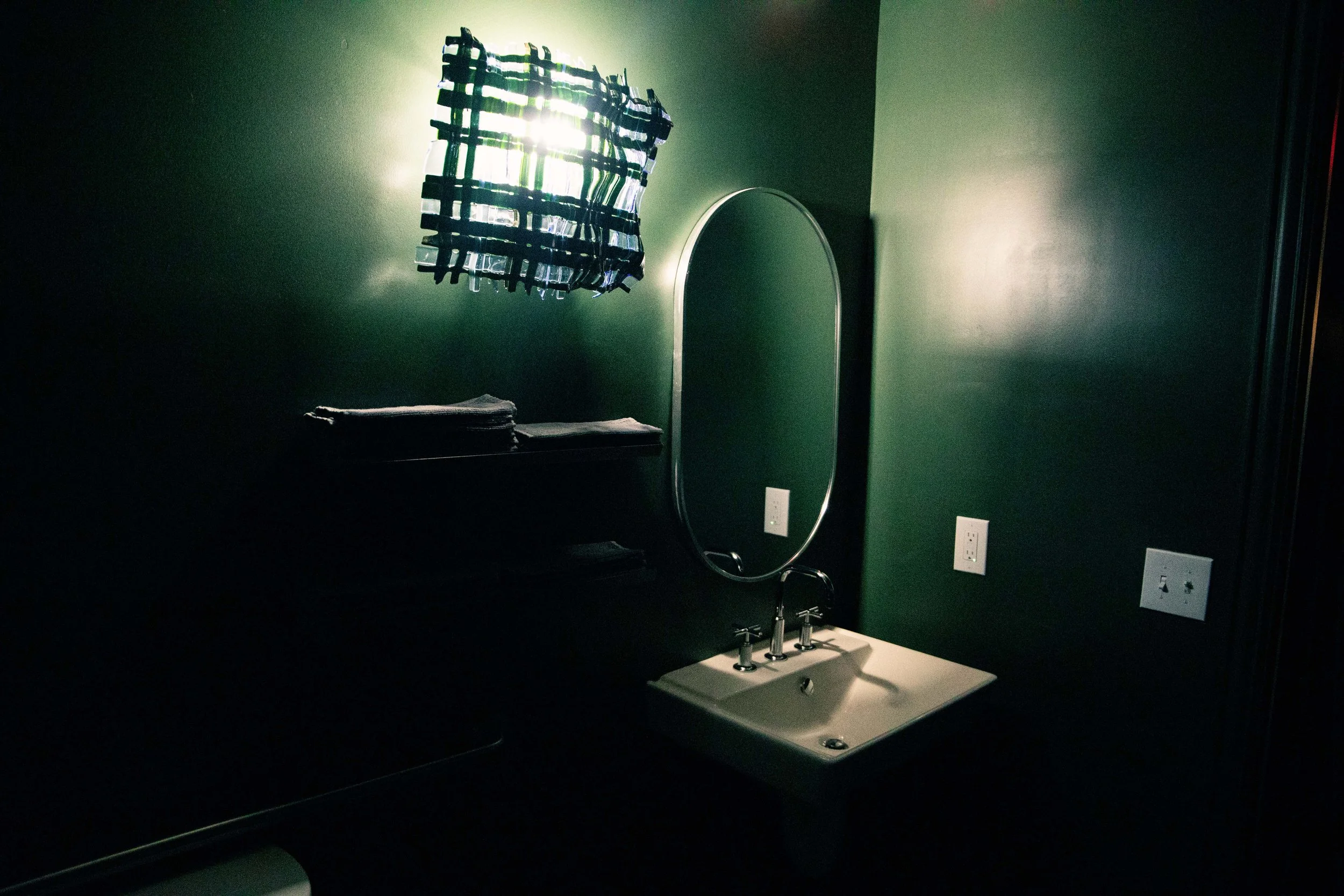 A bathroom with a green wall, a mirror, a sink, and a window with a blue plaid curtain. The mirror is oval, and the sink has two faucets. There are folded towels on a shelf and electrical outlets on the wall.